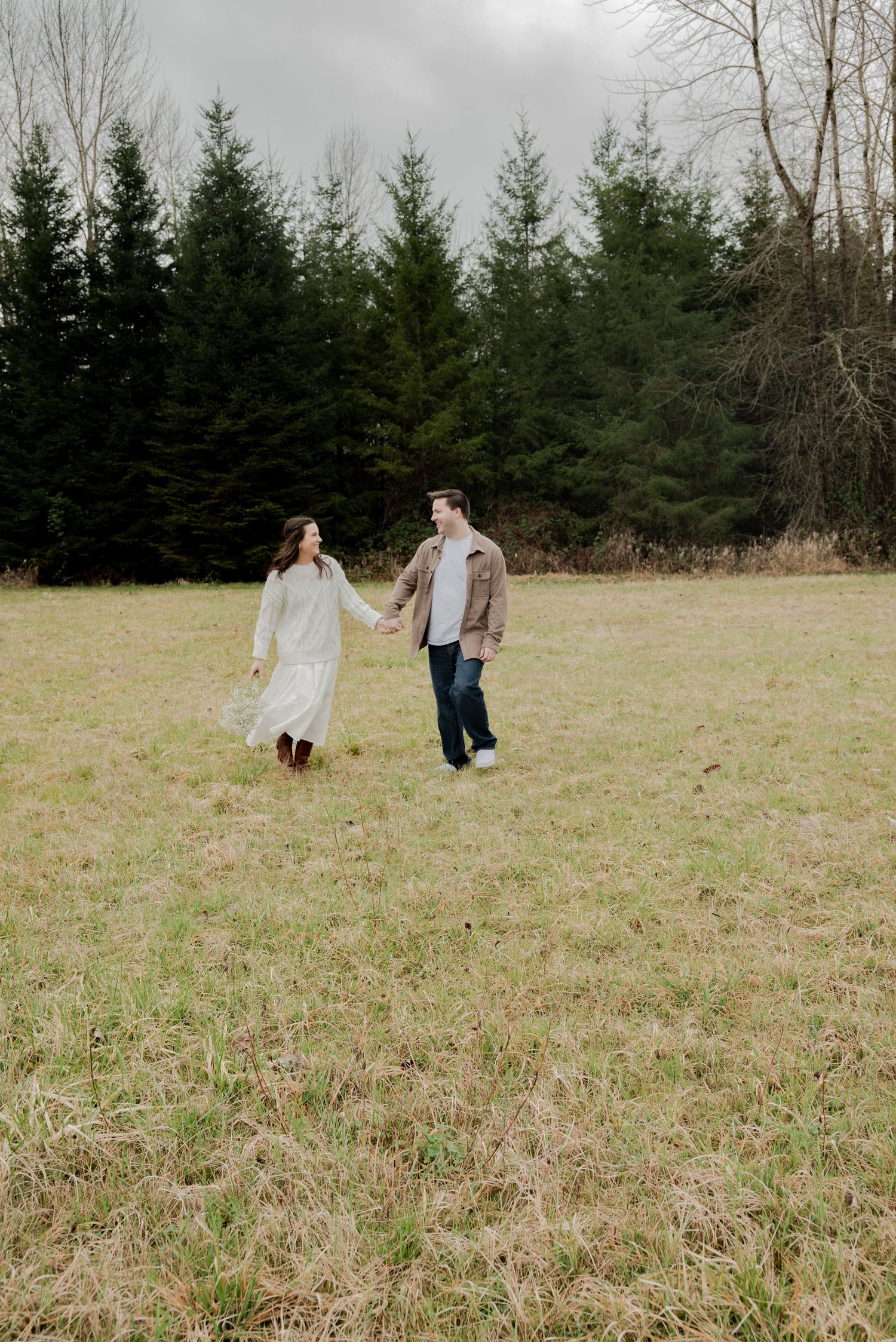 Deming, Washington, A couple walks together in a grassy field bordered by evergreen trees on an overcast day.