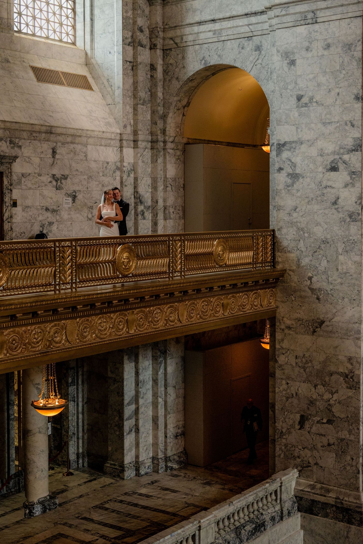 Bride and groom standing on an ornate golden balcony inside a grand marble building interior.