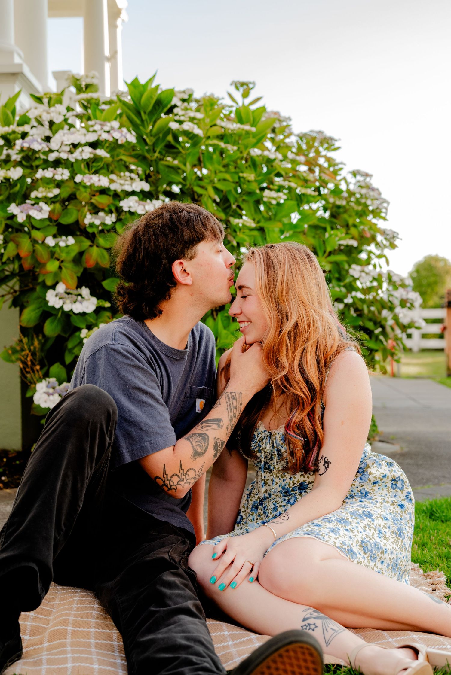 Young tattooed couple sharing a tender kiss while sitting on a blanket in front of flowering bushes.