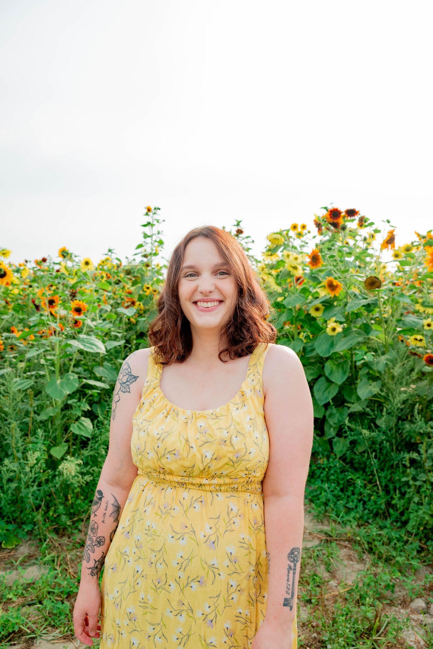 A person in a yellow floral dress stands smiling in front of a field of blooming sunflowers on a sunny day.