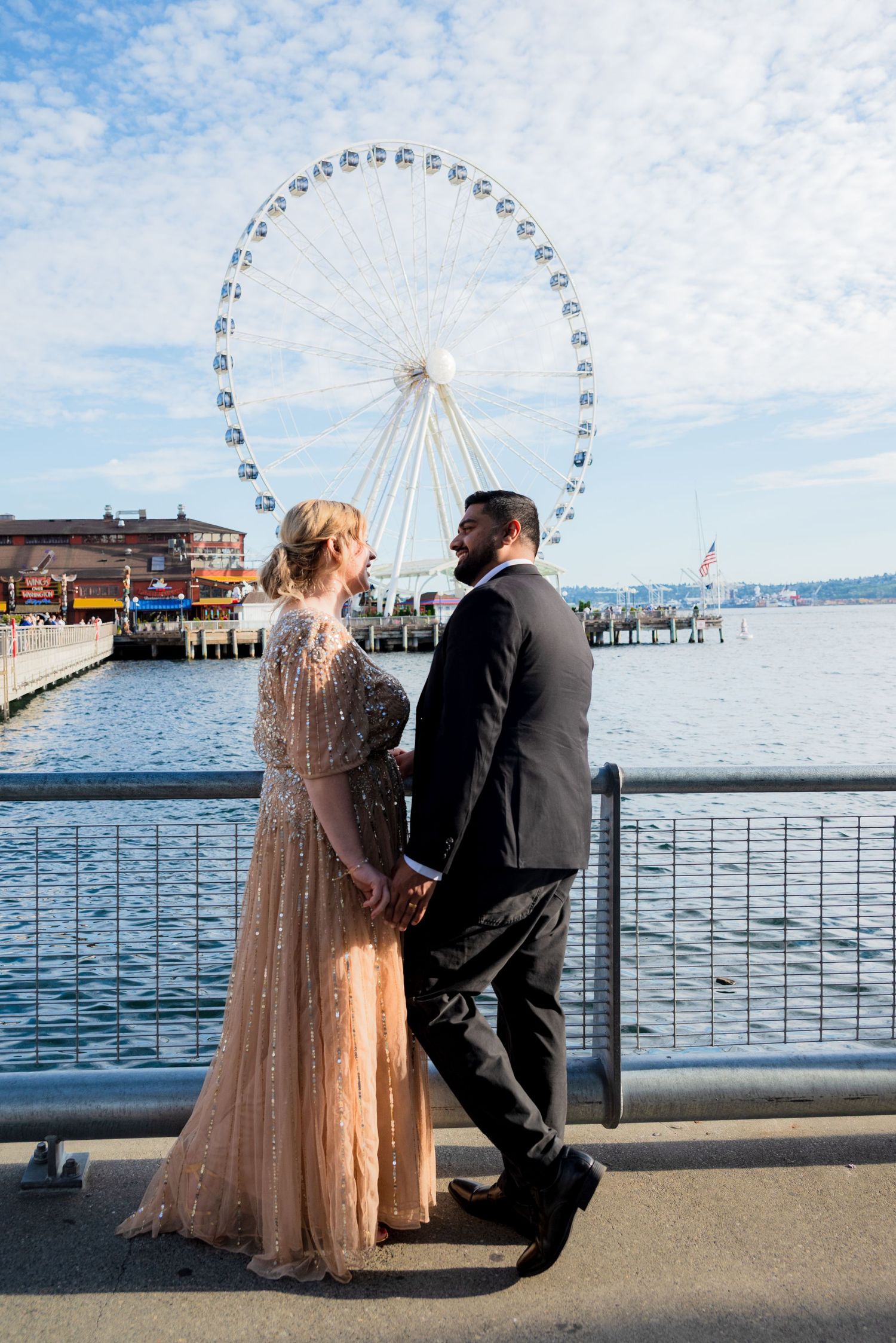 Couple holds hands facing Seattle Great Wheel ferris wheel on waterfront under cloudy blue sky.