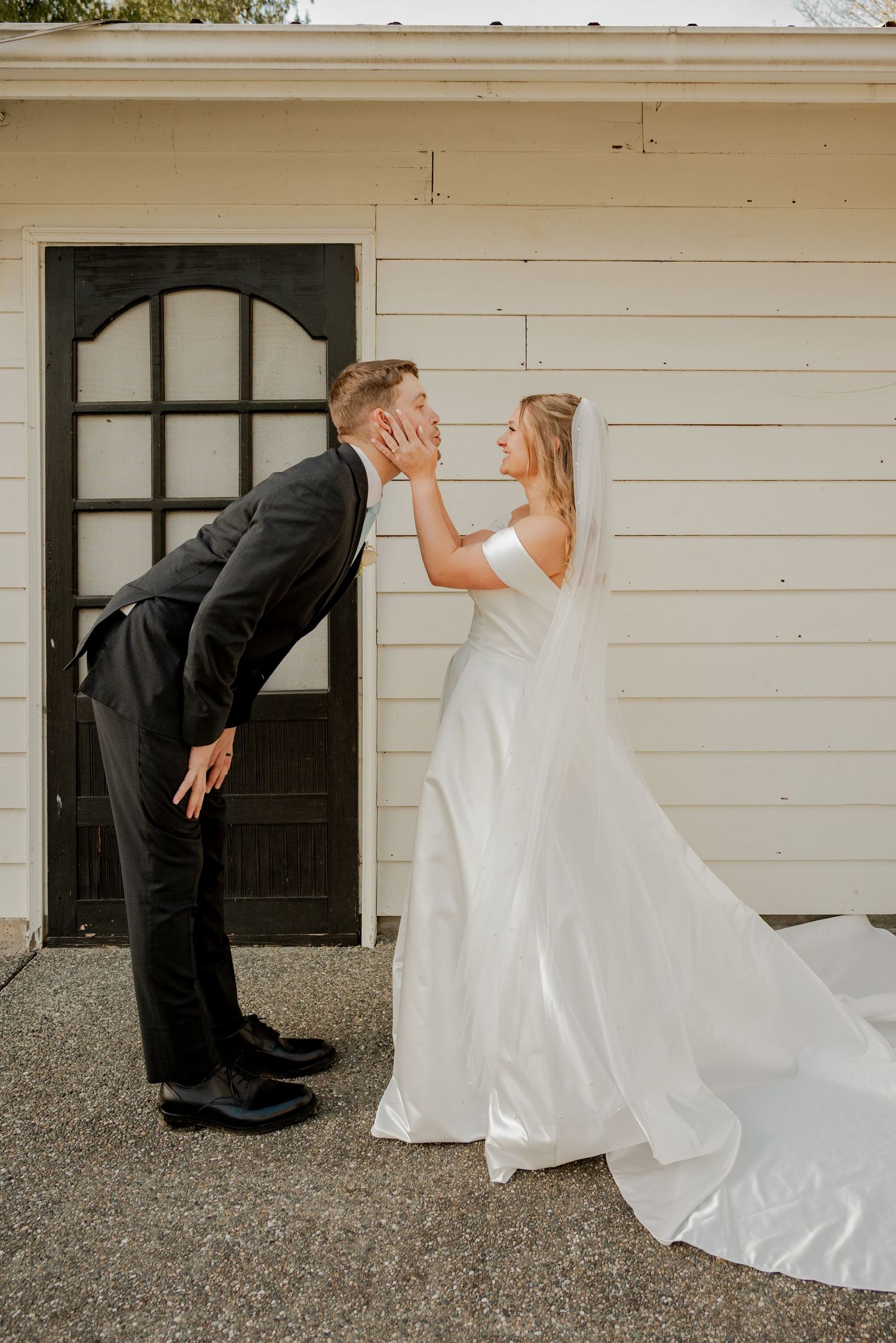 Tall groom in black suit leans down to kiss blonde bride in white gown outside a white building.