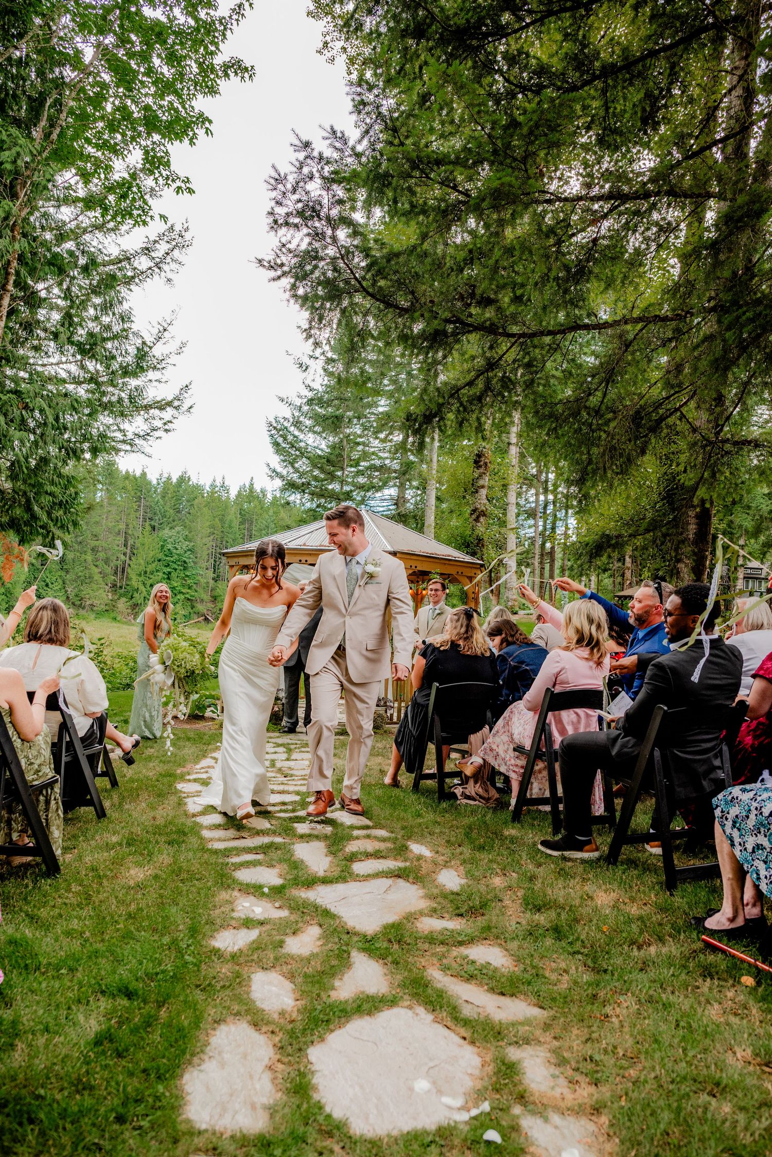 Newlyweds walking down a stone aisle through a forest wedding ceremony as guests celebrate around them.