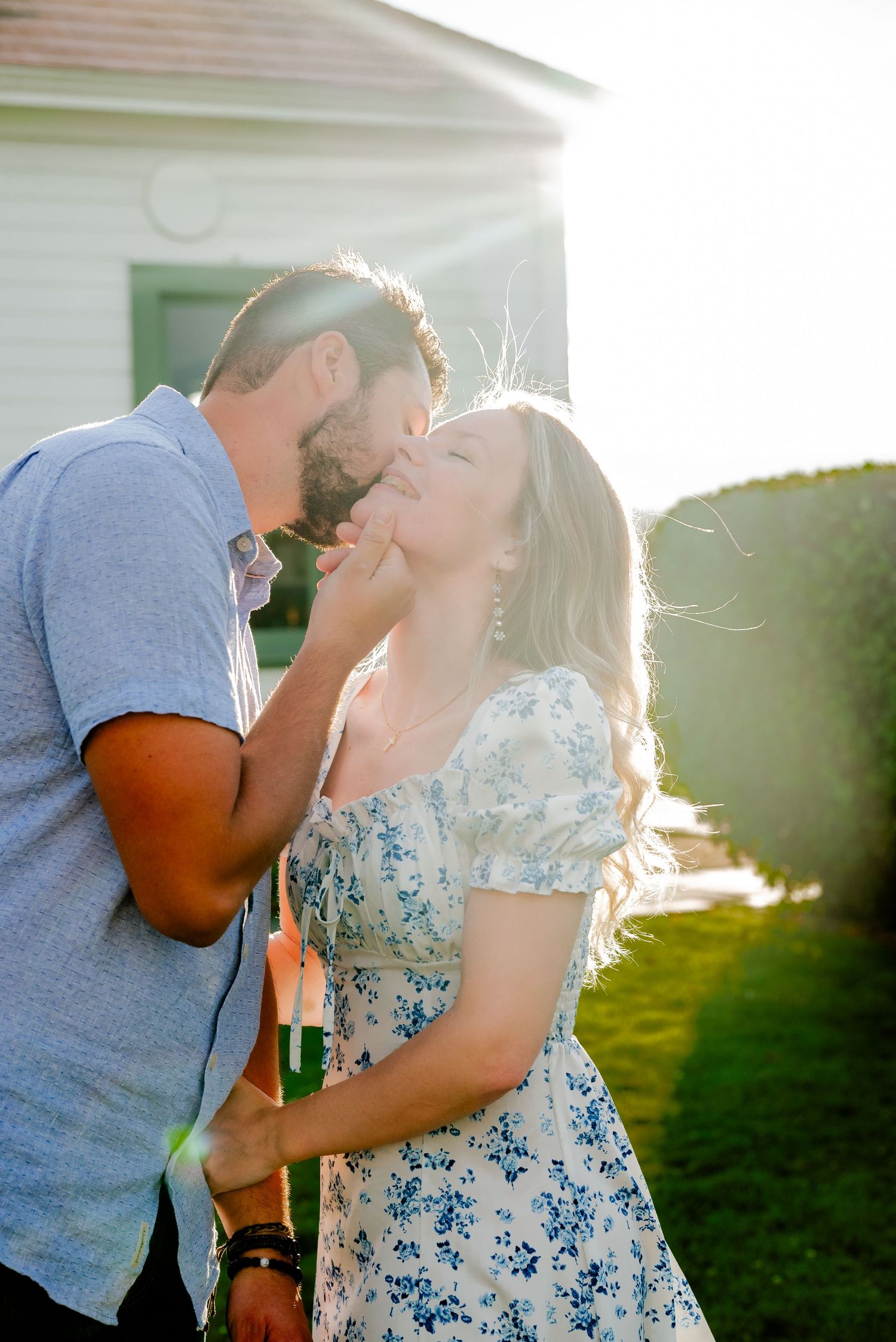 Couple sharing a romantic kiss outdoors in golden sunlight, woman in floral dress, man in blue shirt, lush greenery behind them.