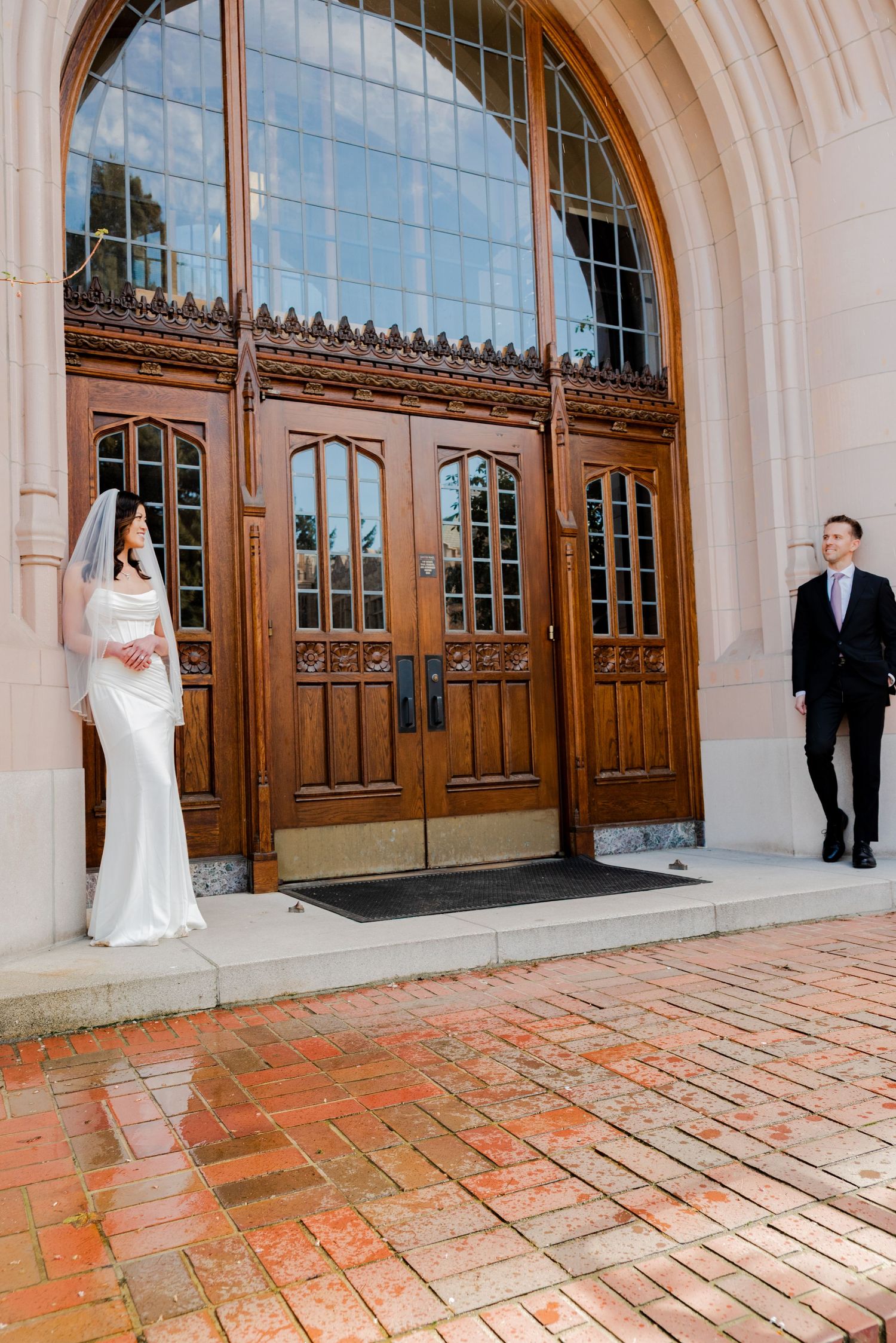 Seattle Washington, A bride in white gown and groom in tuxedo stand on opposite sides of grand wooden church doors with arched architecture.