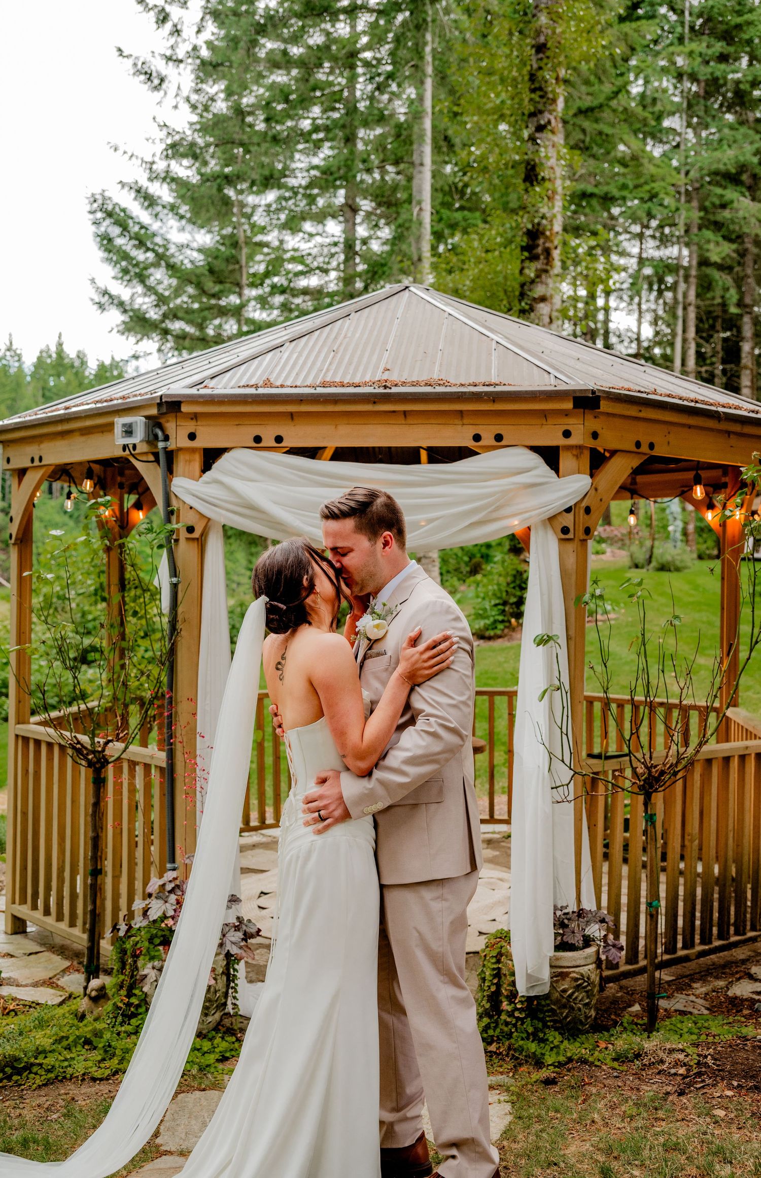 Bride and groom share first kiss under decorated wooden gazebo surrounded by tall evergreen trees.