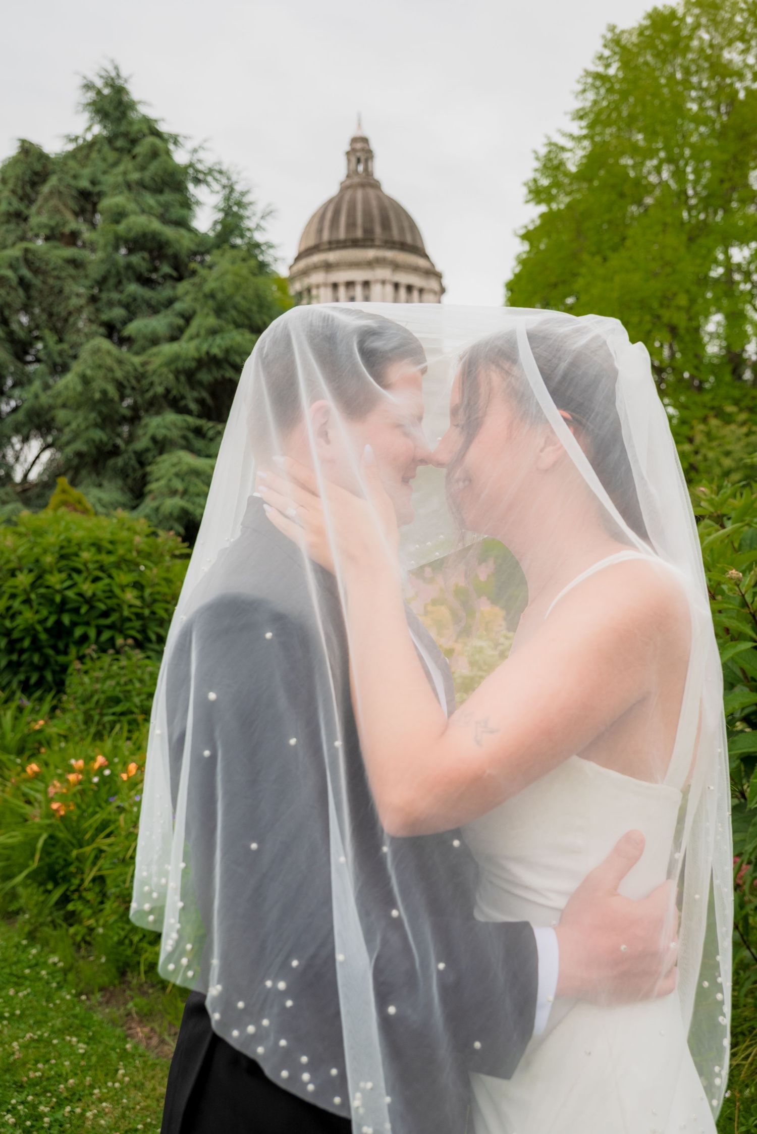 Two brides share a romantic kiss under a flowing veil with a domed capitol building in the background.