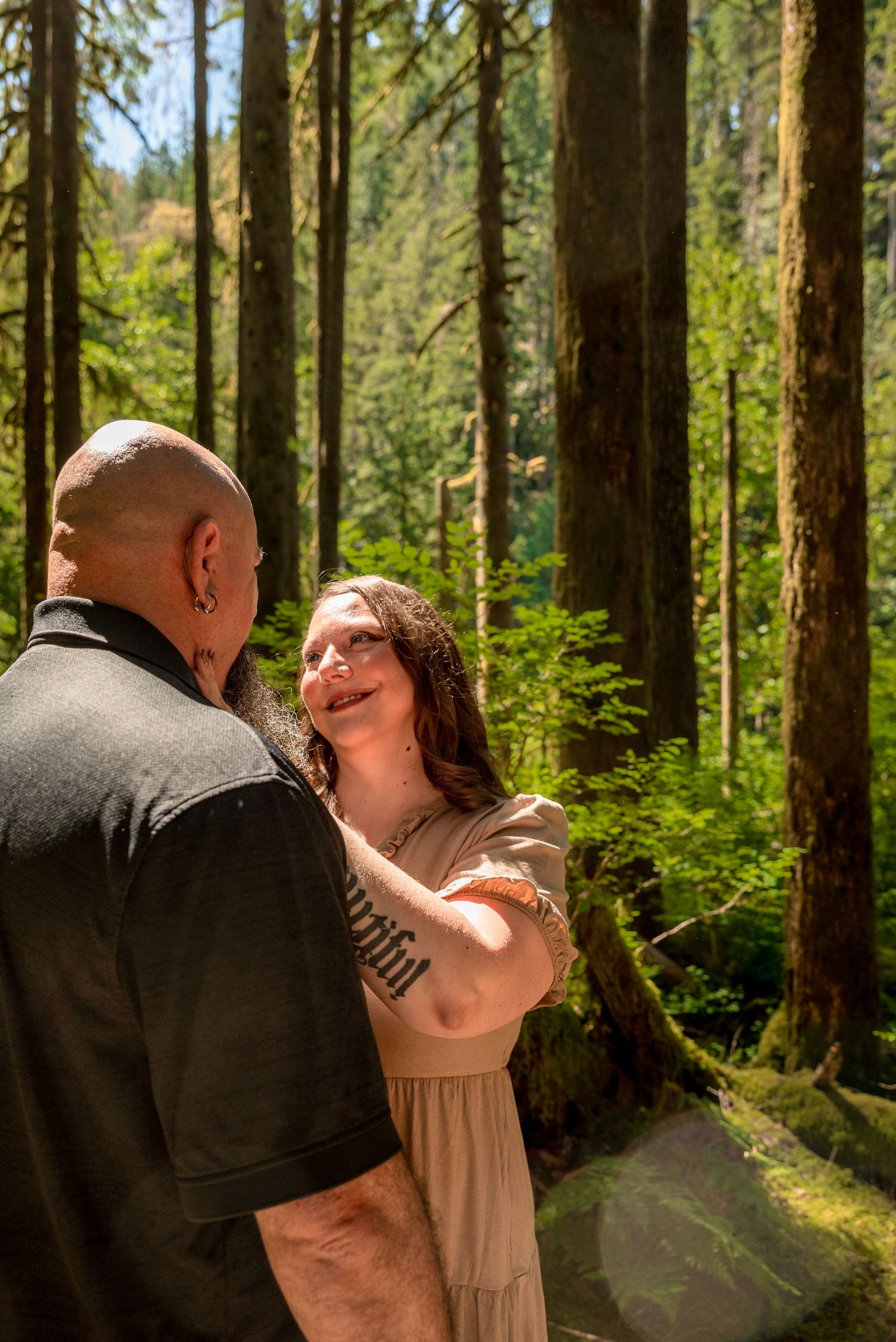 Couple sharing a romantic moment in a lush green forest, woman gazing up at bald man among tall mossy trees.