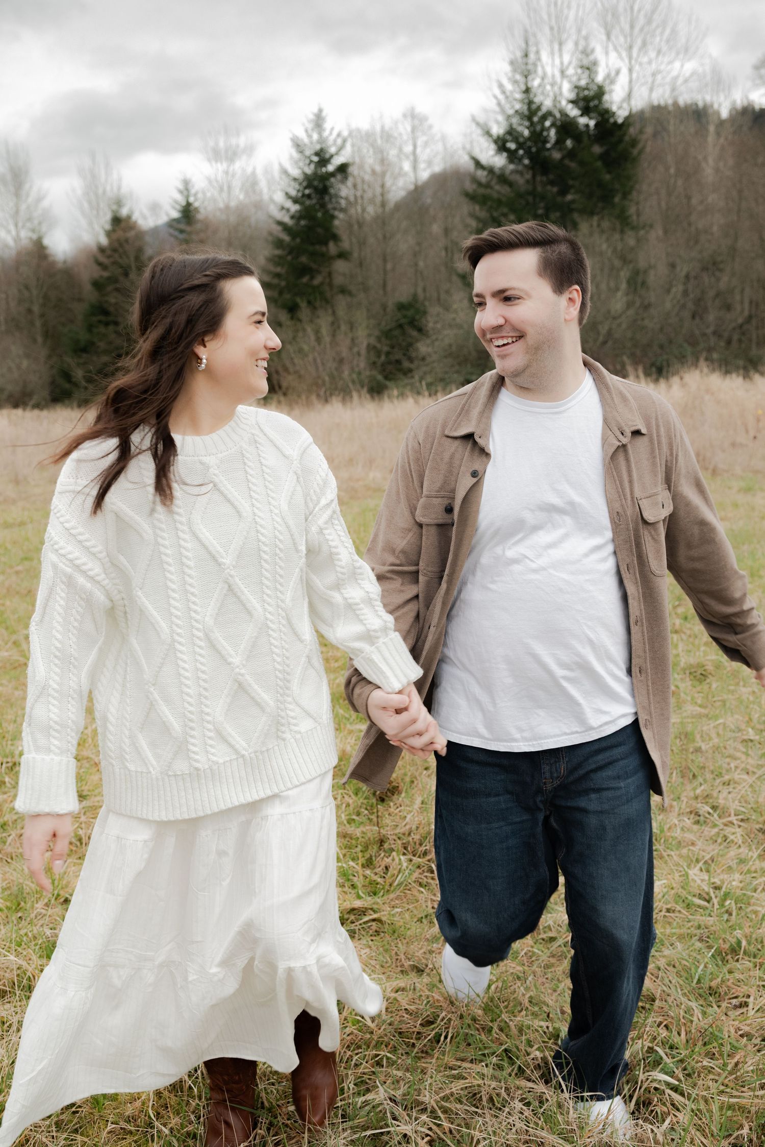 Deming, Washington, A couple holding hands and laughing together in a field with evergreen trees in the background on an overcast day.