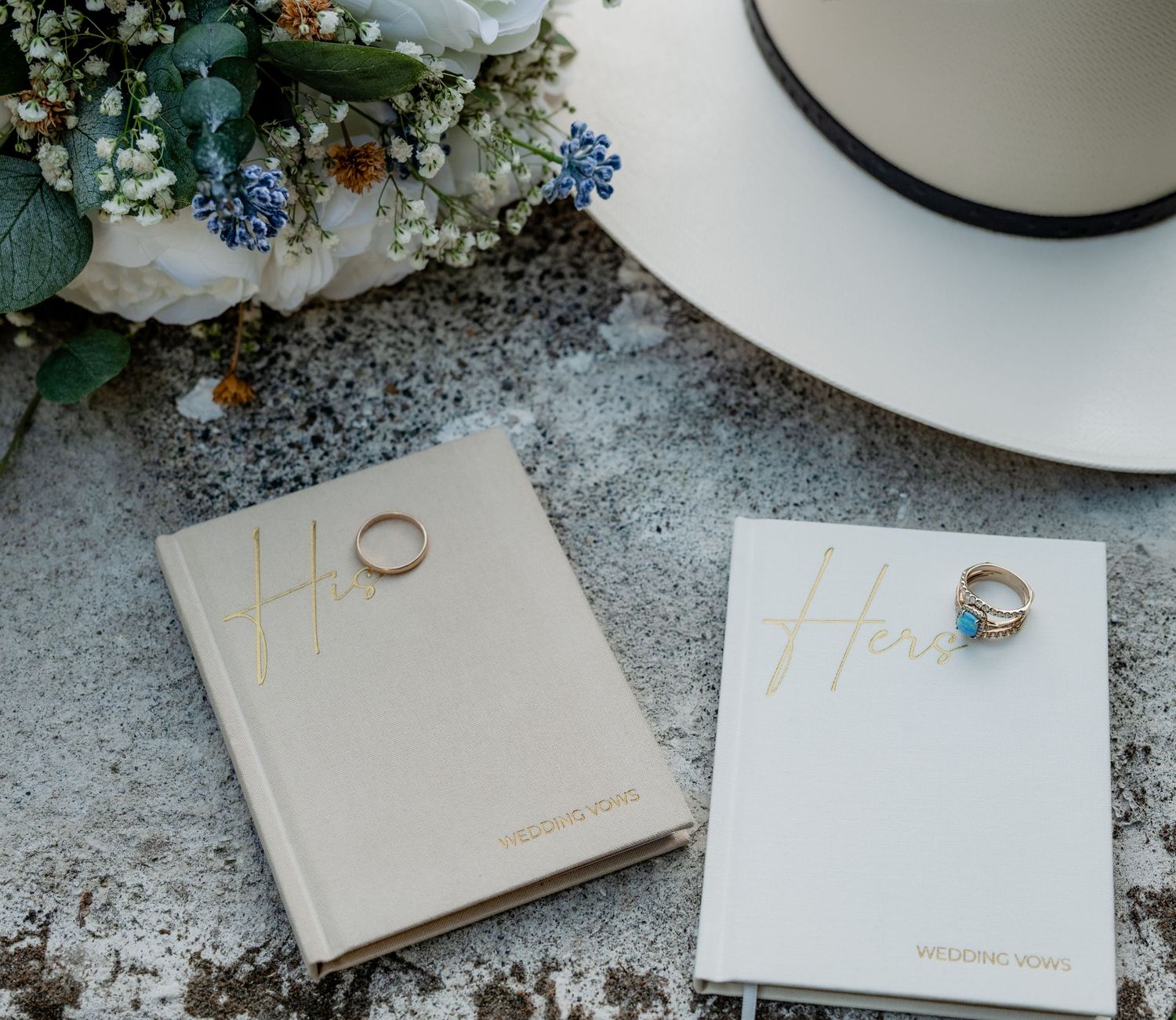 Two wedding vow books labeled Her with gold rings and a white hat beside a floral bouquet on a marble surface.