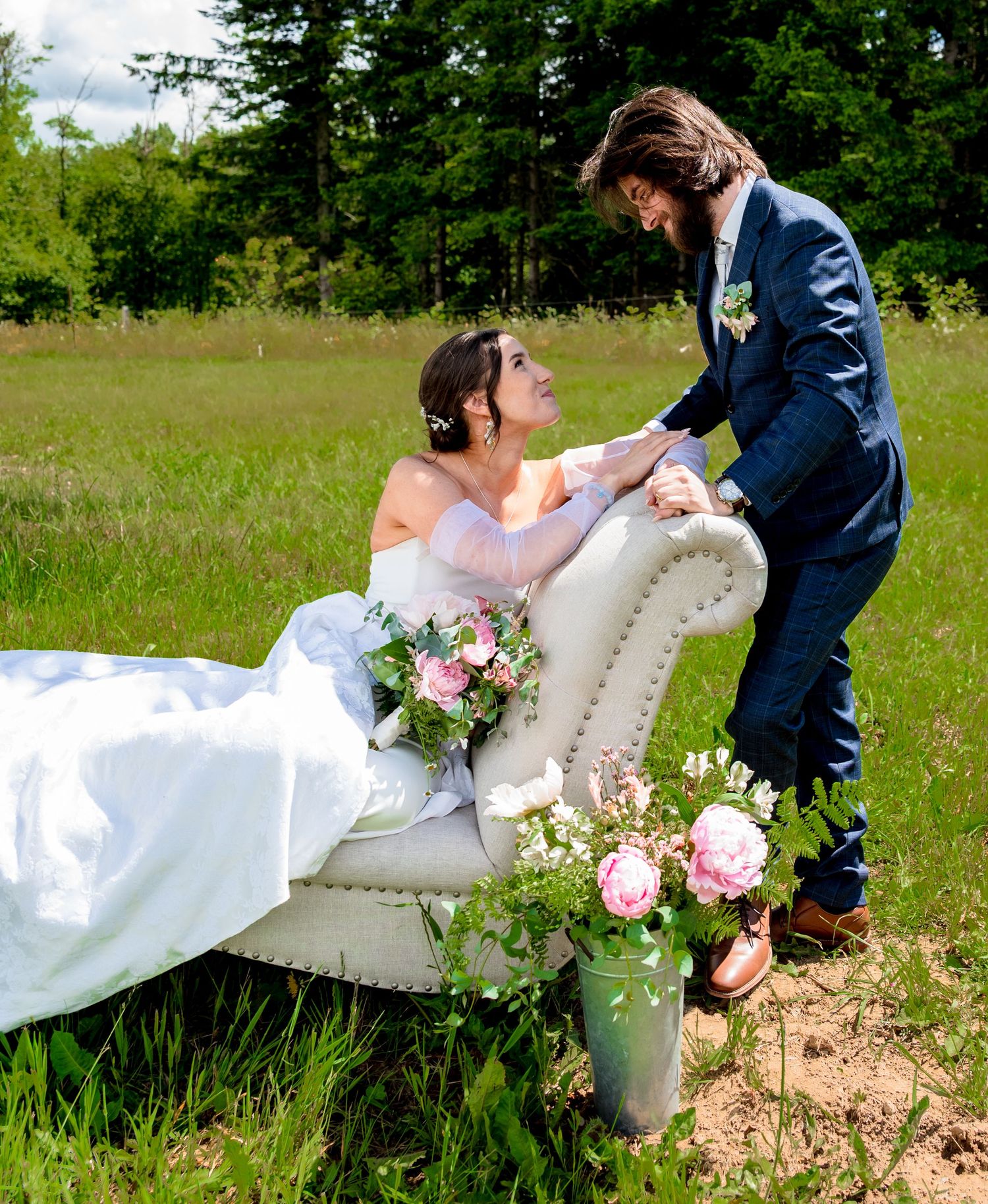 Bride in white gown reclines on vintage chair as groom in navy suit holds her hands in a lush meadow.