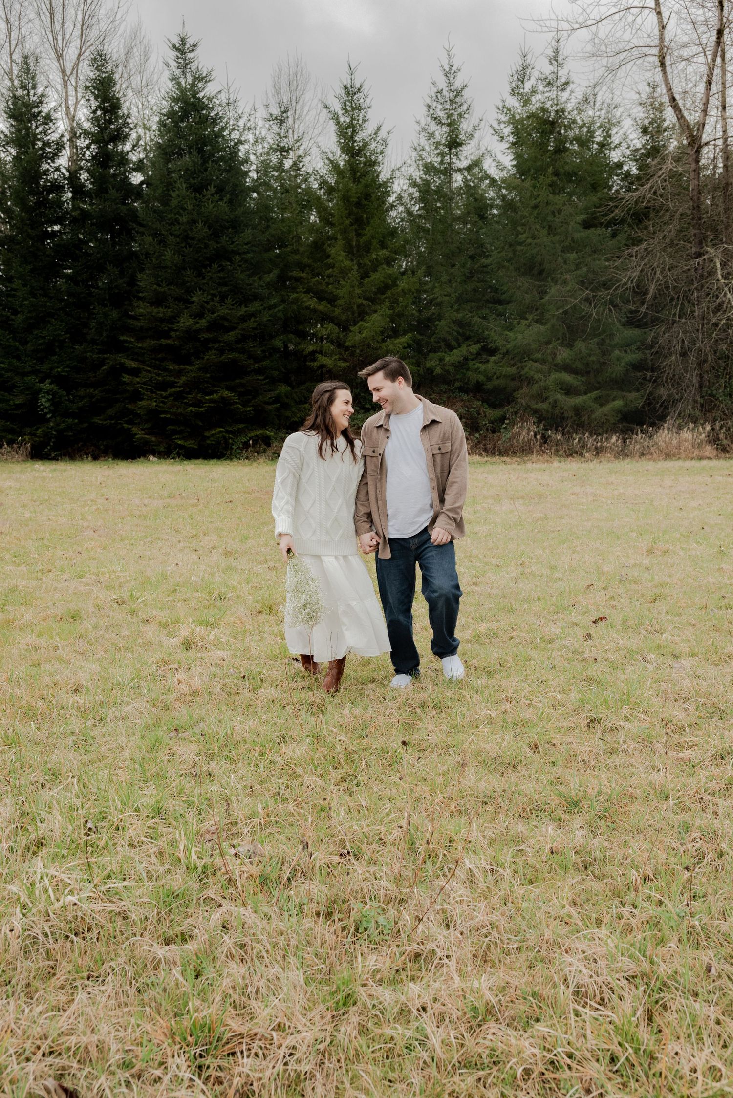 Deming, Washington, A couple walks together through a grassy field with evergreen trees in the background on an overcast day.