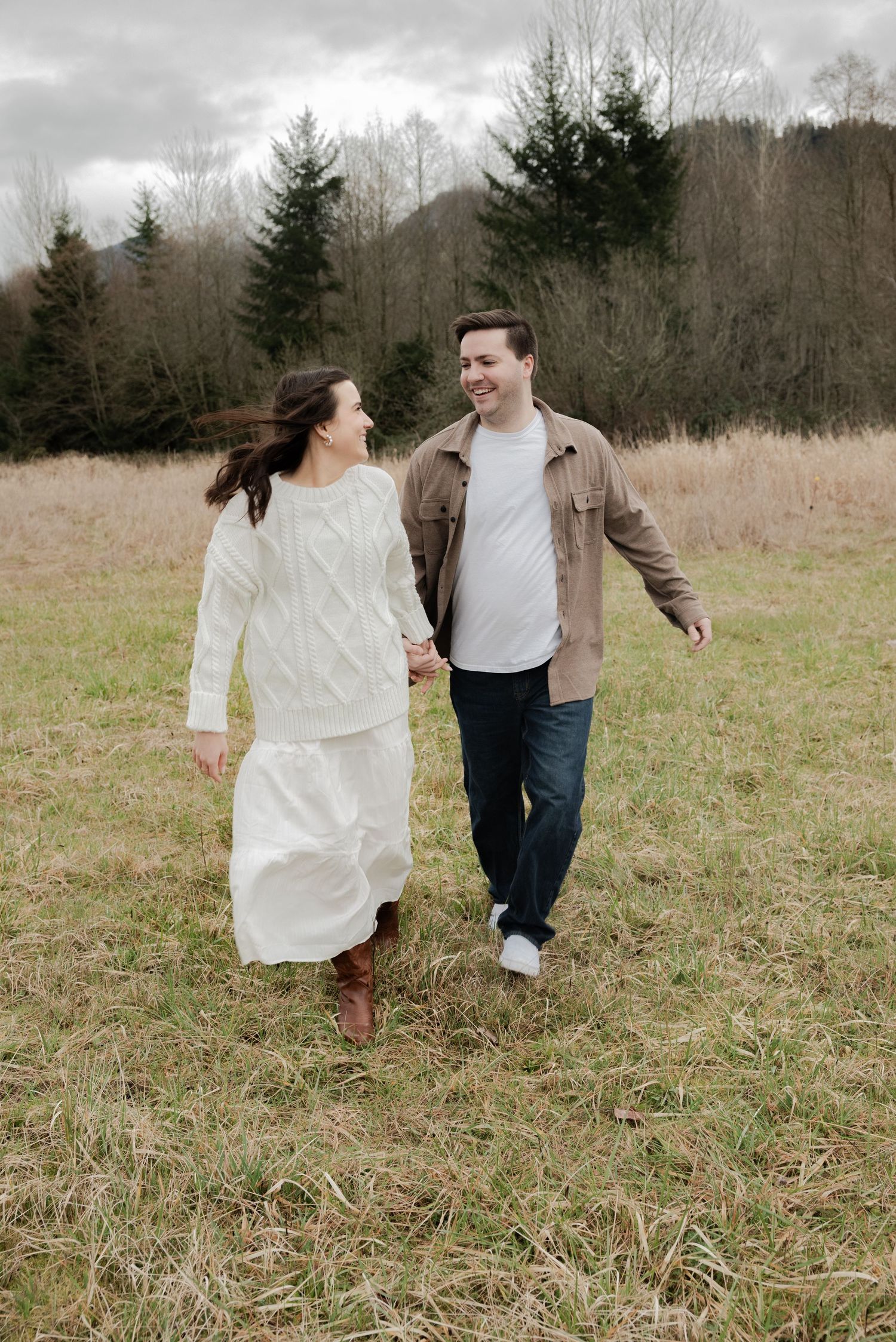 Smiling couple walking hand in hand through an open field with trees and cloudy skies behind them.