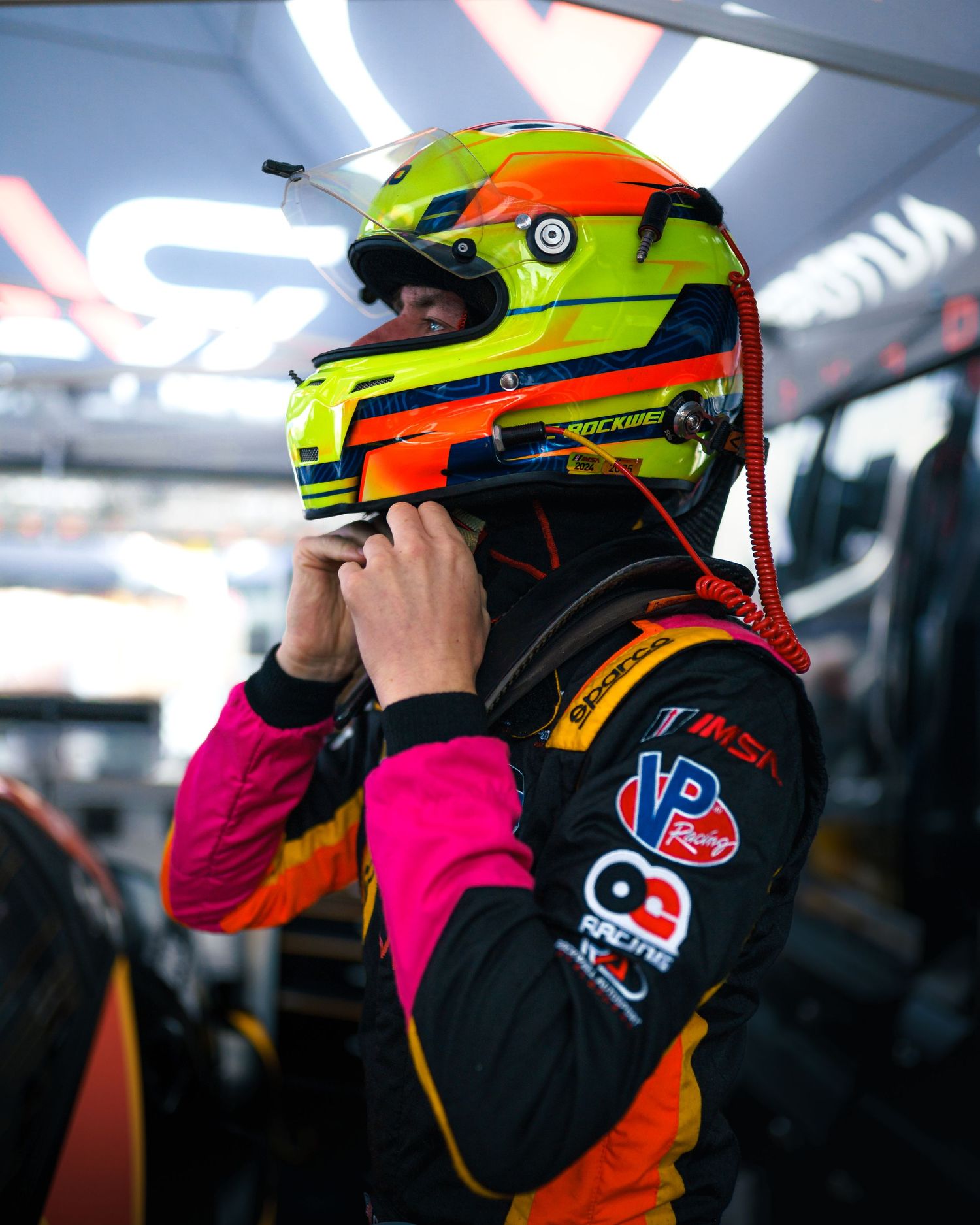 Race car driver adjusts neon yellow and orange helmet while wearing black and pink racing suit with sponsor patches.