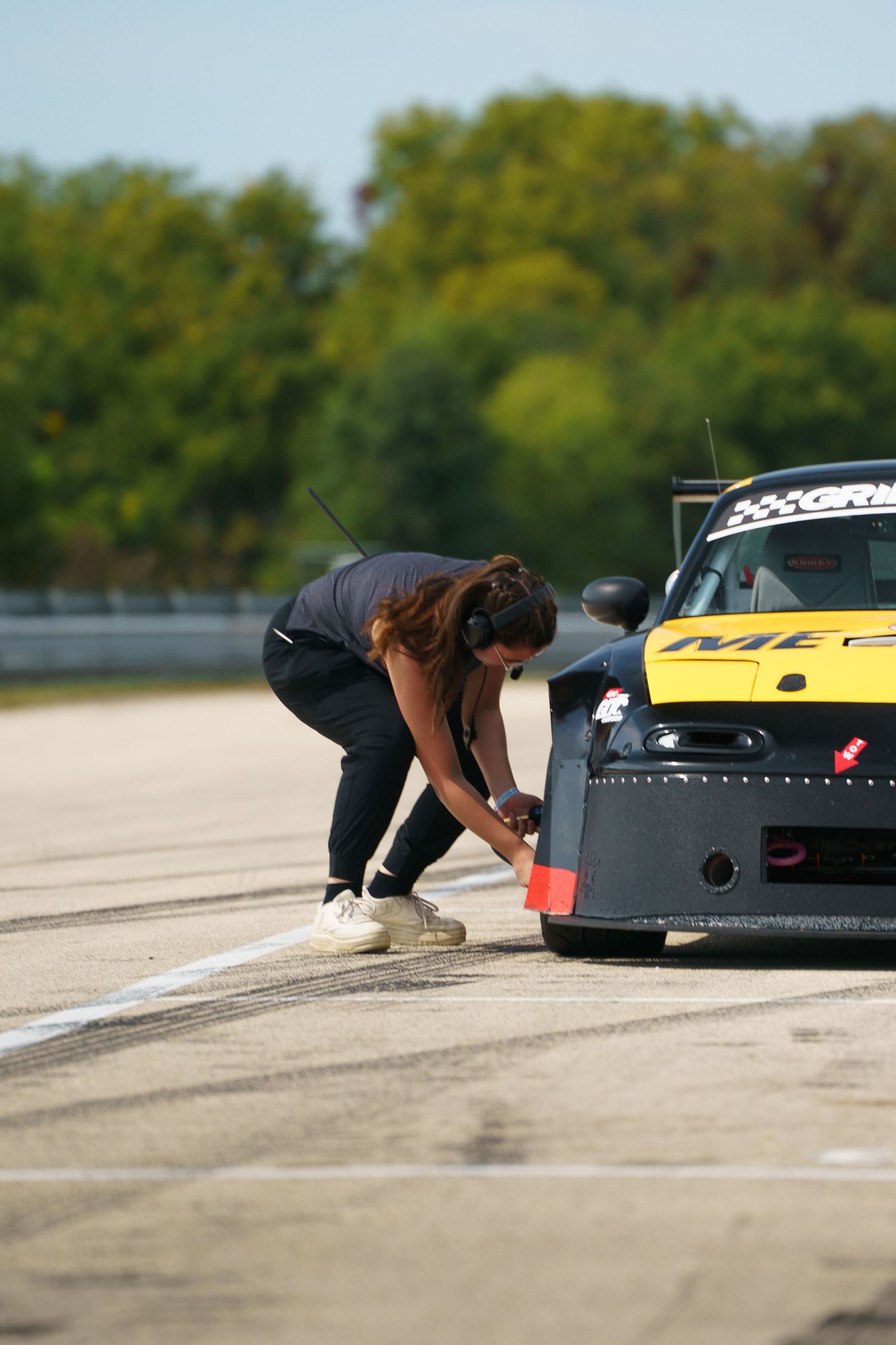 Race crew member bends down to inspect a yellow and black race car on a track with green trees in background.