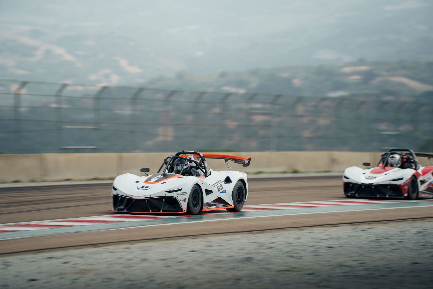 Formula race cars compete on a racetrack with mountains visible in the background during a motorsport event.
