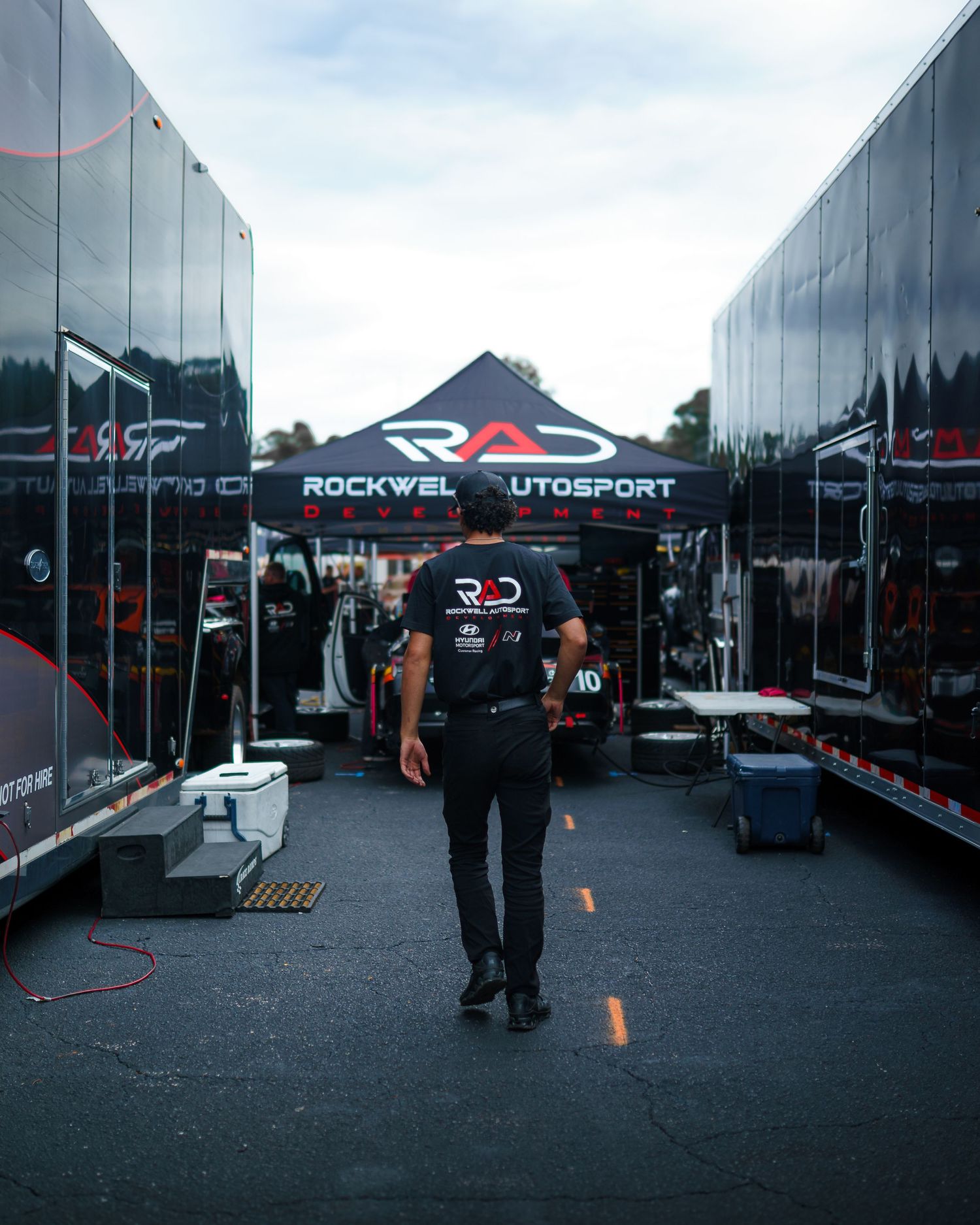 Racing team transporters and pit equipment lined up in the paddock area at a motorsport event.