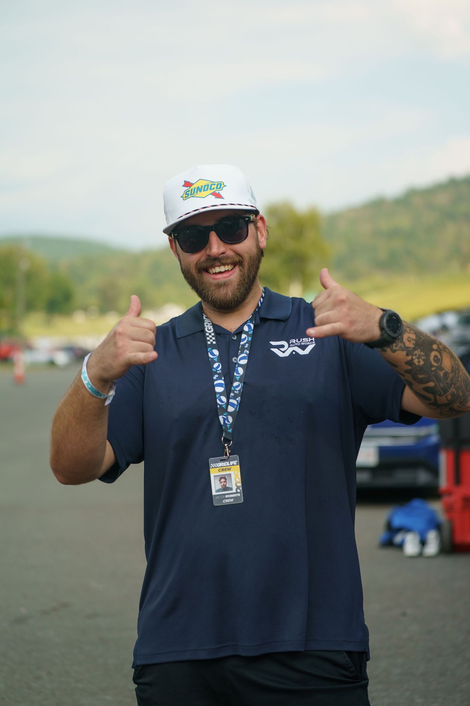 A person in a navy blue polo shirt and baseball cap gives thumbs up gestures while standing outdoors.
