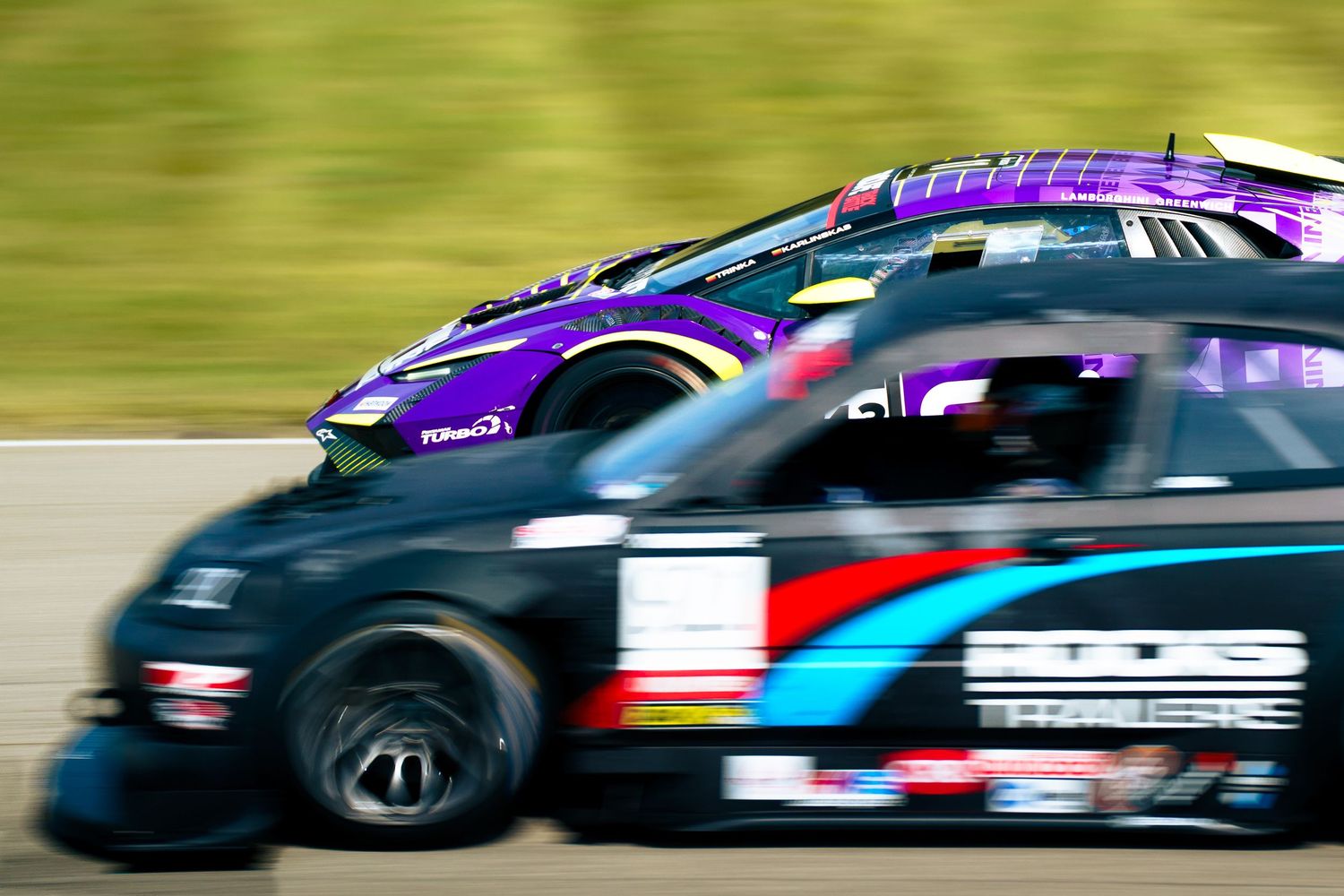 Racing cars compete at high speed with motion blur showing purple and black vehicles on a racetrack.
