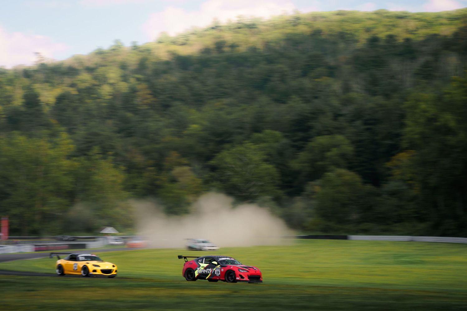 Race cars speed down a track surrounded by lush green trees and dust clouds as they compete for position.