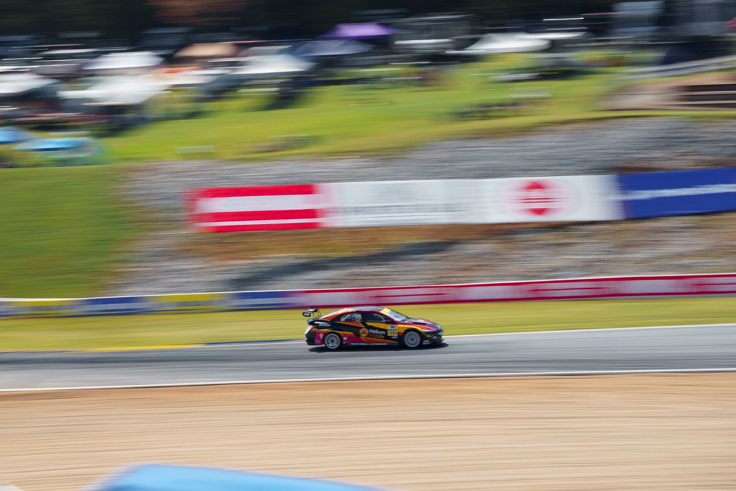 Red race car speeds through a racing circuit with green grass and spectator areas visible in the background.