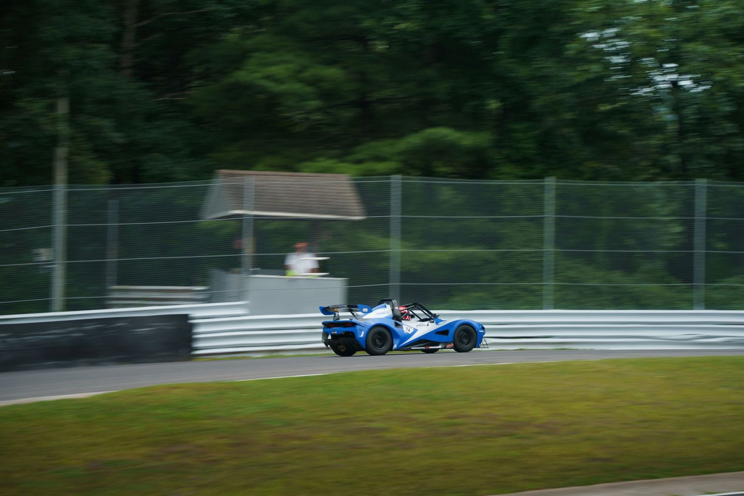 Race car speeds through a forested racetrack with other cars visible in the distance during a motorsport competition.