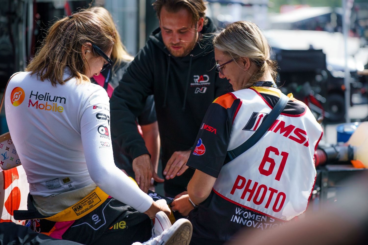 Racing pit crew members gather around a car during a motorsport event while wearing branded uniforms.