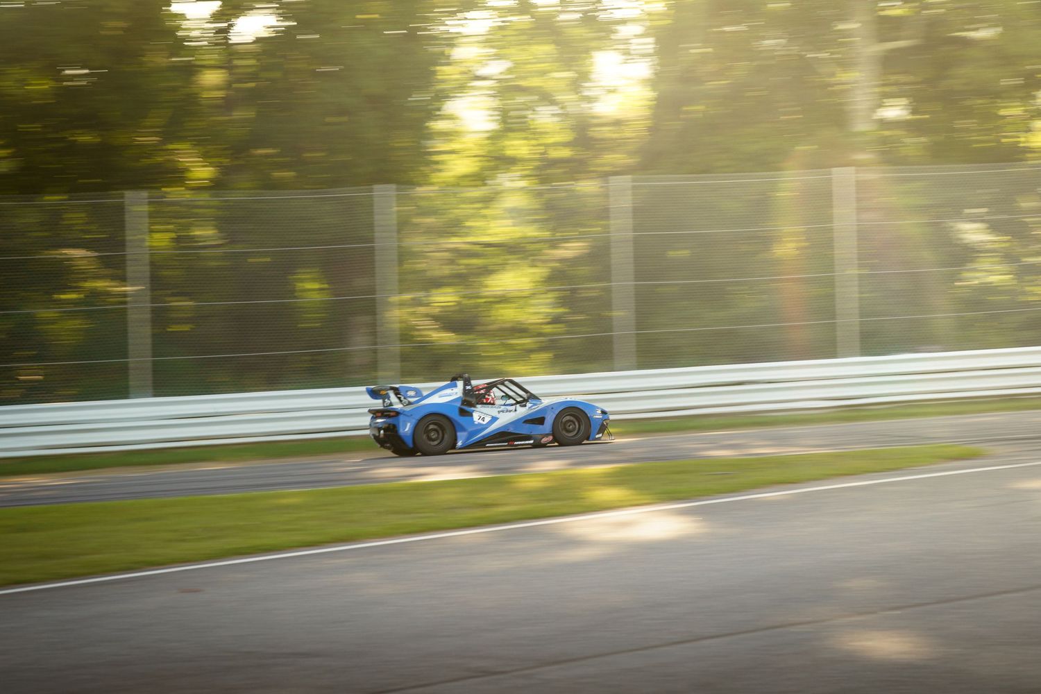 A blue race car speeds through a sunlit forested racetrack with rays of light filtering through the trees.