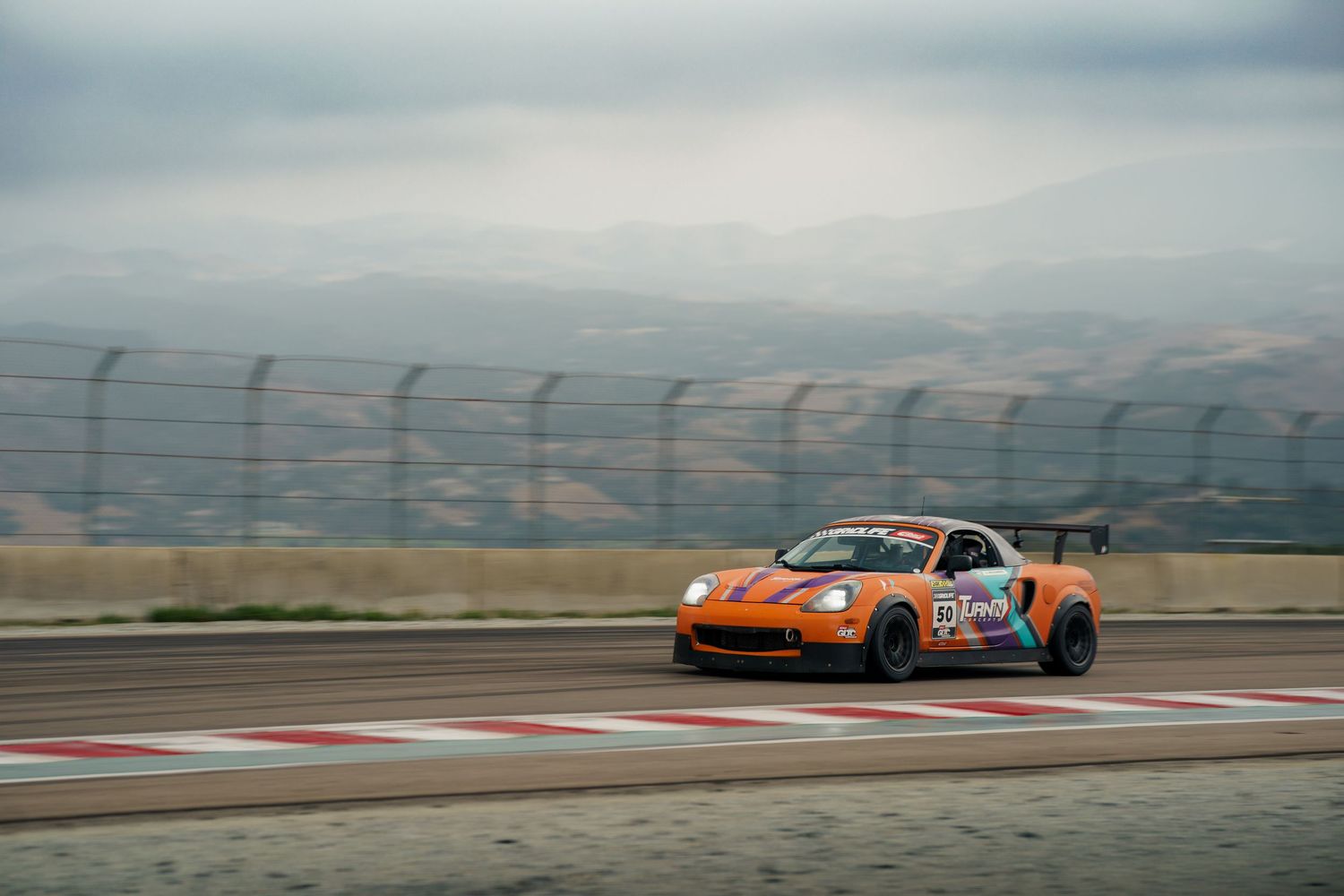 Racing cars speed around a winding track with mountains in the background during a competitive motorsport event.