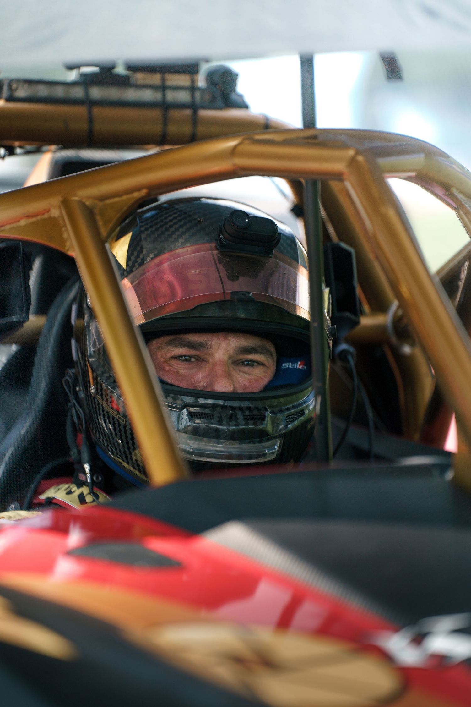 Racing driver in helmet visible through roll cage of competition vehicle.
