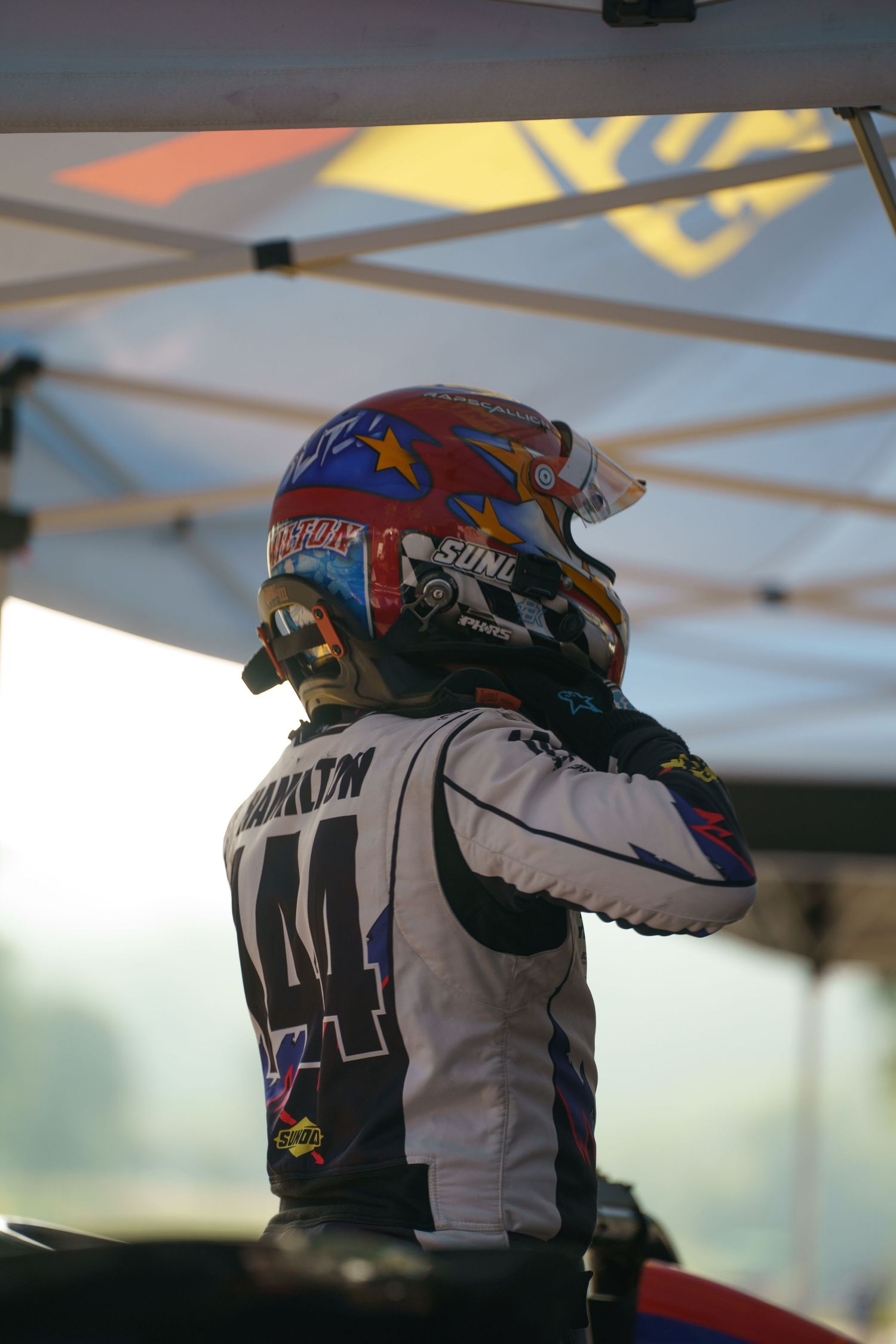 Racing driver in a white jersey with number 44 wearing a colorful helmet is sitting at the track.