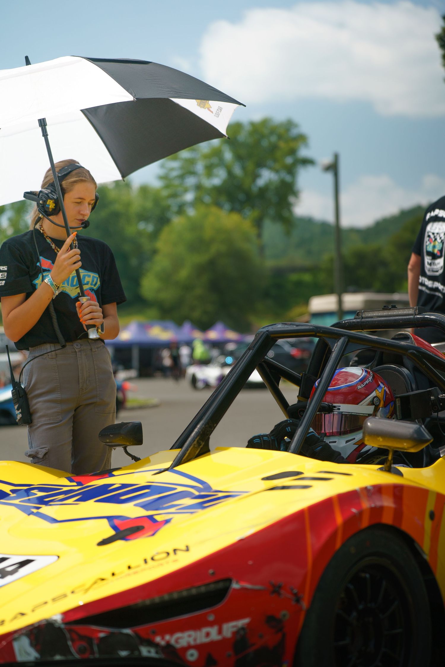 A person holds a black umbrella while photographing a yellow and red race car on a sunny day at a motorsports event.