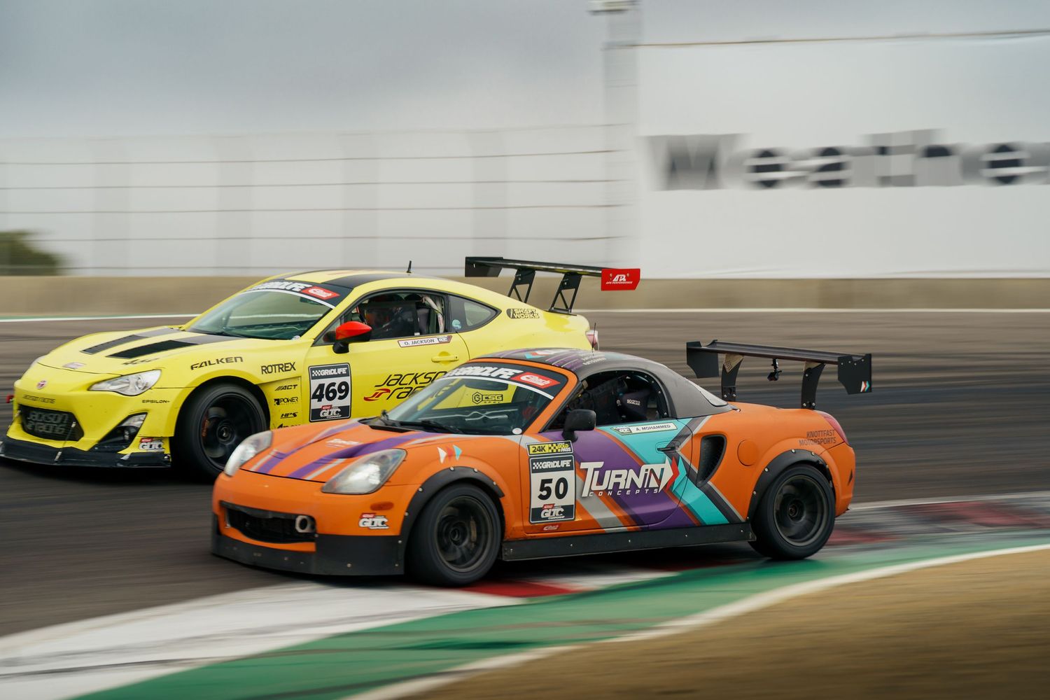 Colorful race cars speed around a racetrack corner during a competitive motorsport event.