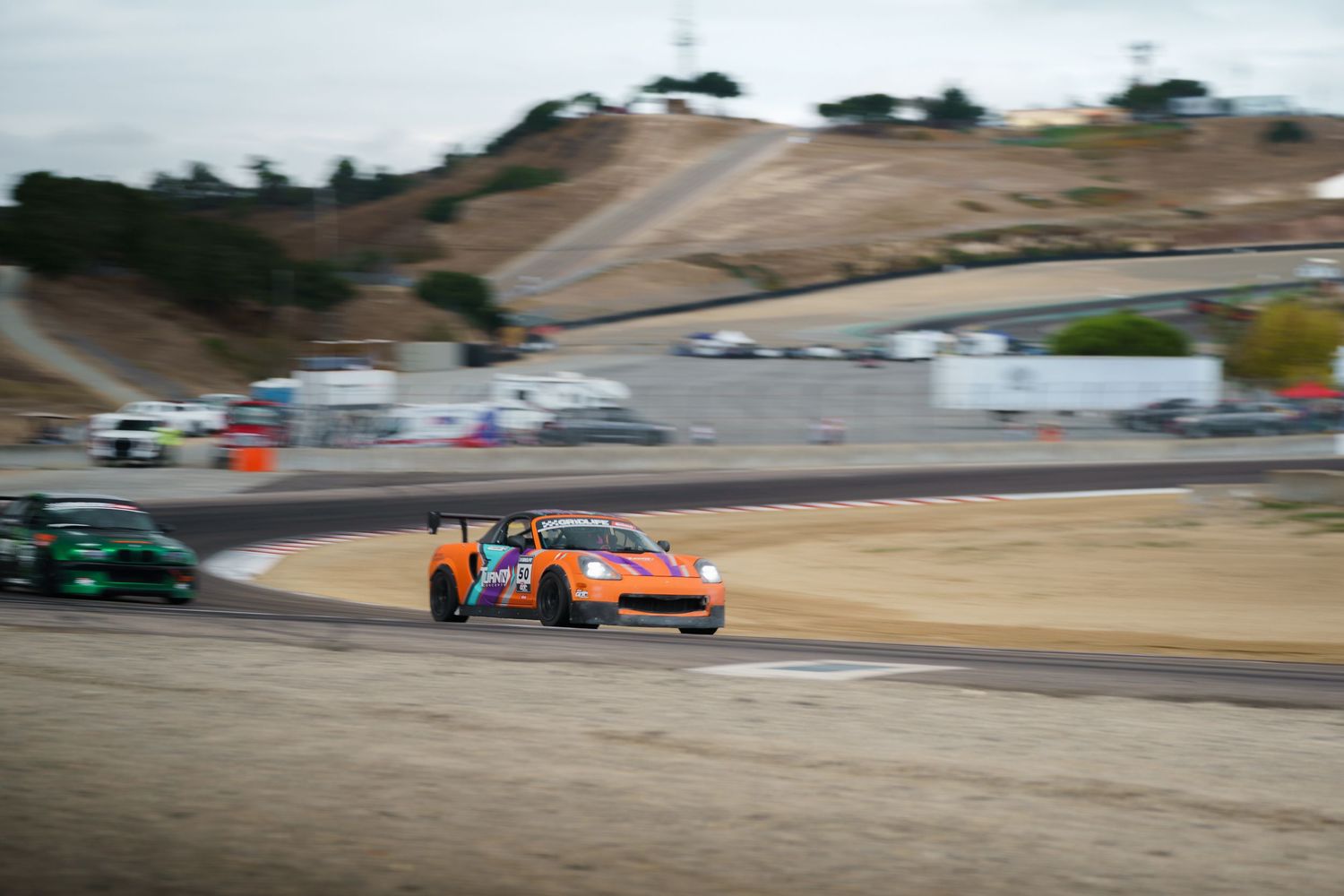 Orange race car speeds through a turn on a race track with rolling hills in the background.