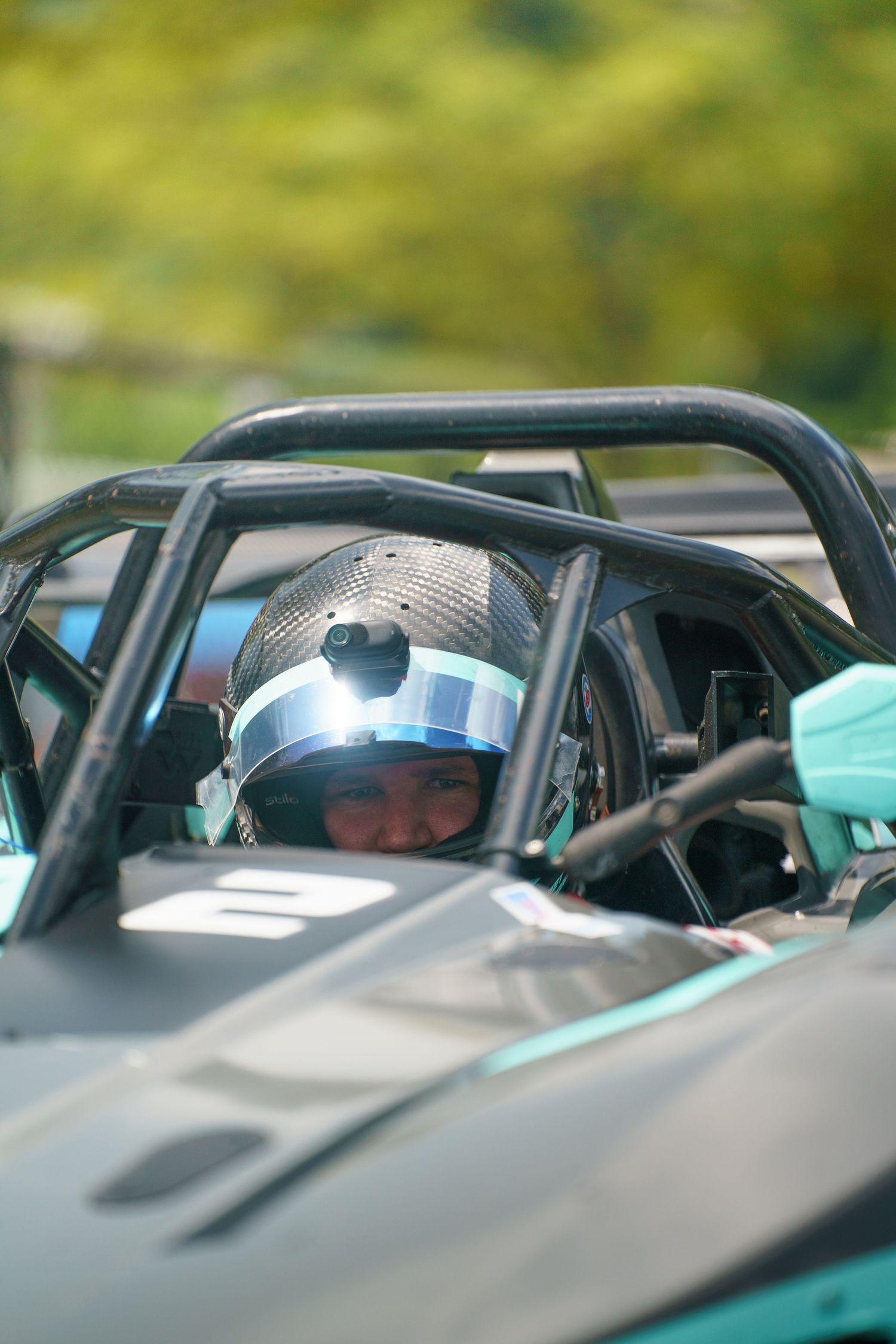 A race car driver in a helmet sits inside the roll cage of a sleek black and turquoise racing vehicle.