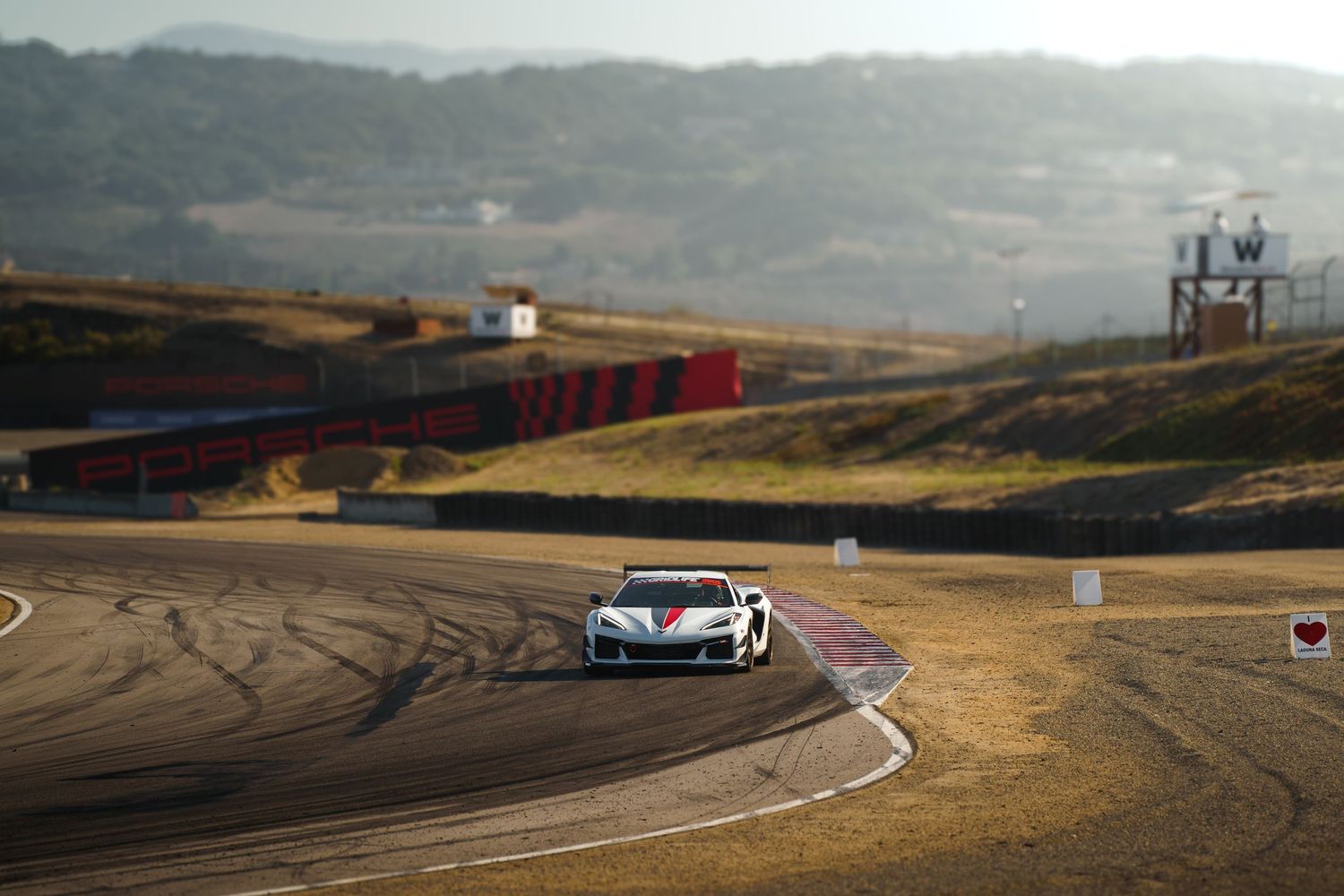 Multiple race cars navigate a challenging turn on a dusty racetrack with rolling hills in the background.