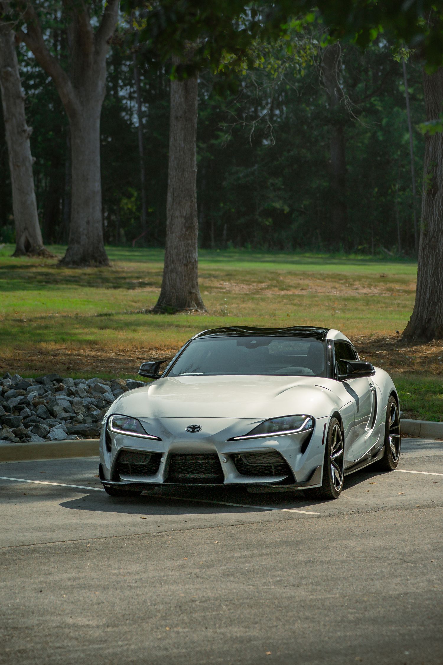 Three-quarter front view of a silver Toyota Supra parked in a shaded wooded area.