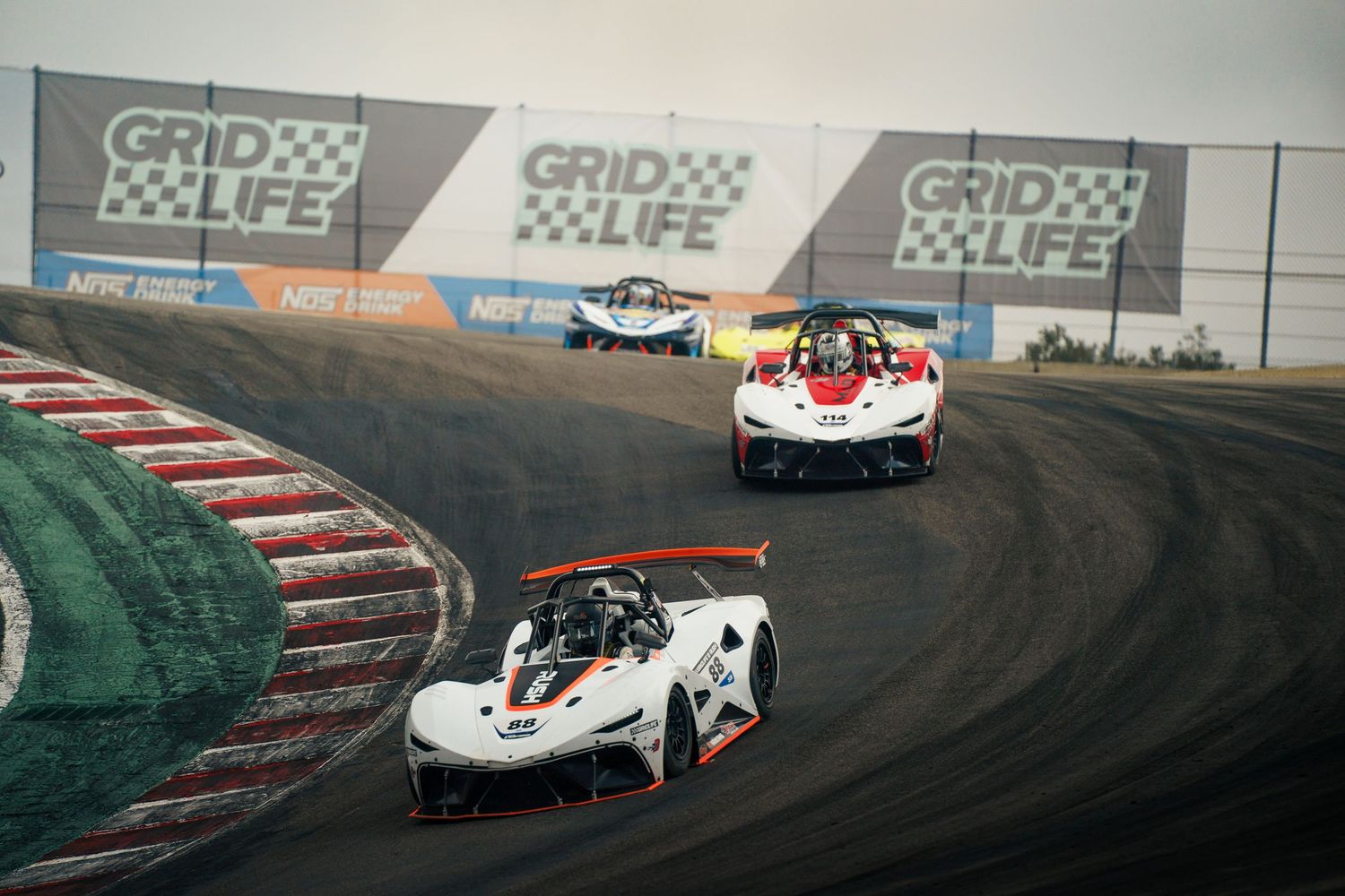 Two white racing cars navigate a challenging turn on a professional racetrack with Grid Life banners visible in background.