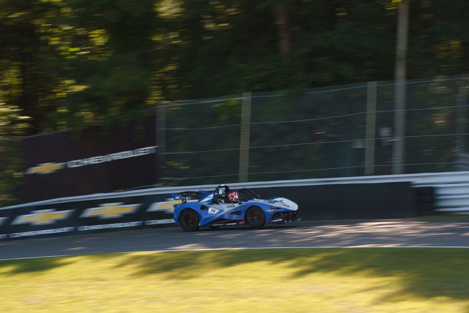 Race car speeds around a sunlit track during a motorsport competition with trees in the background.