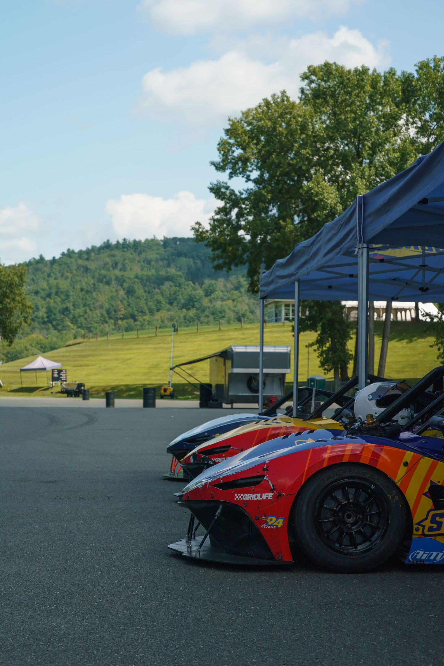 Racing cars are lined up under covered pavilions at a scenic hilltop track on a sunny day.
