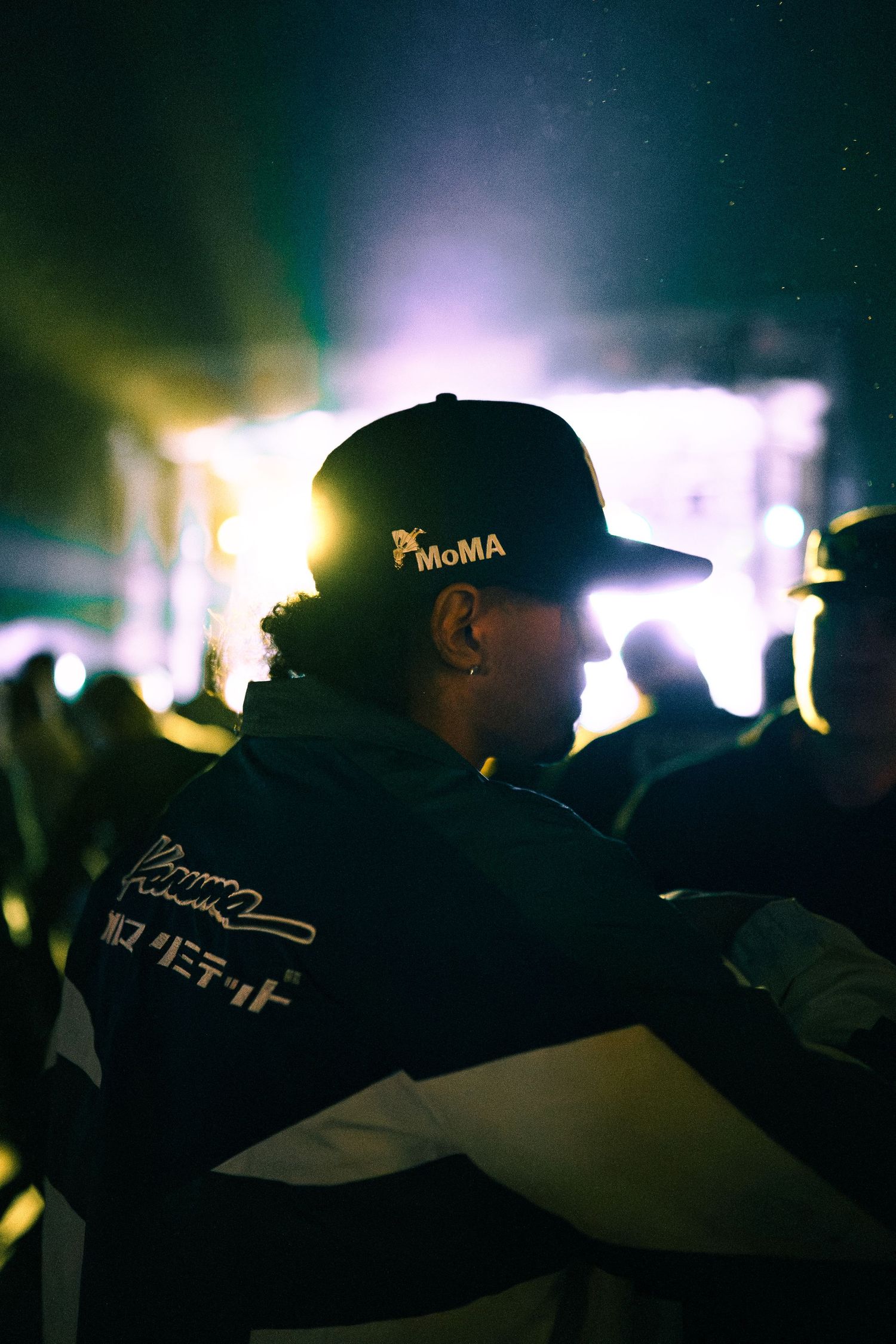 Silhouetted figures in dark baseball caps against blurred concert lighting creating an atmospheric nighttime scene.