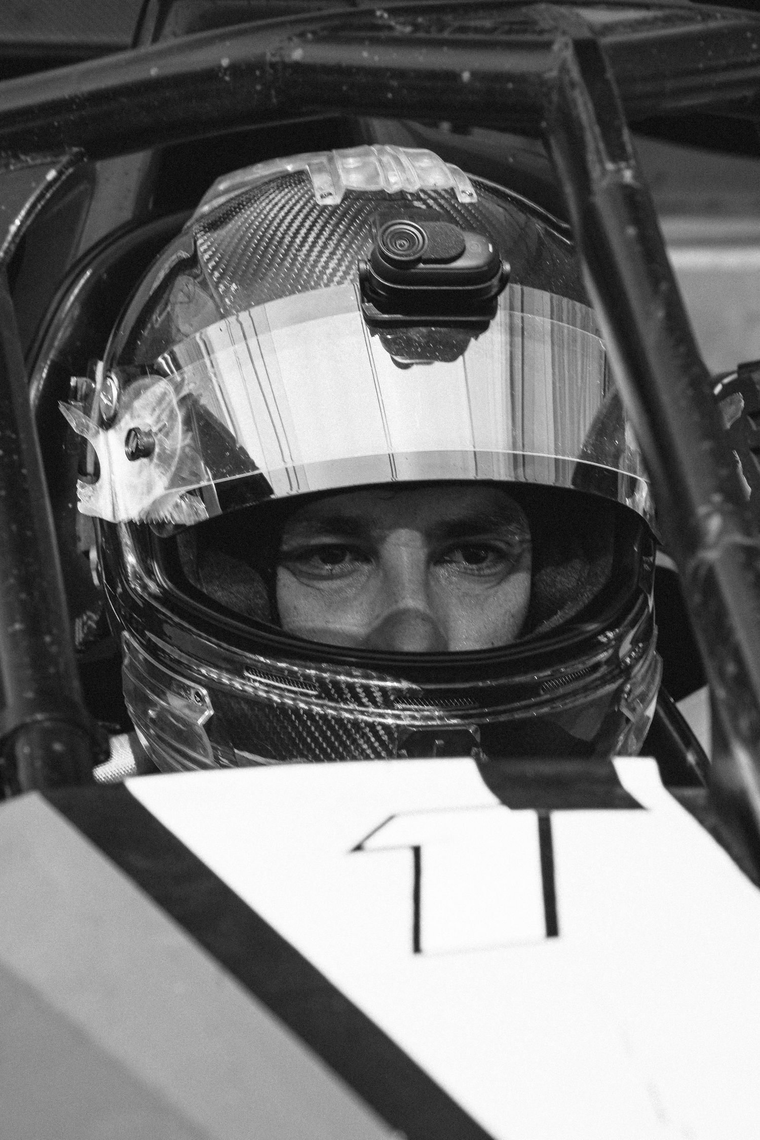 Multiple close-up shots of racing drivers in helmets viewed through car windshields in black and white photography.