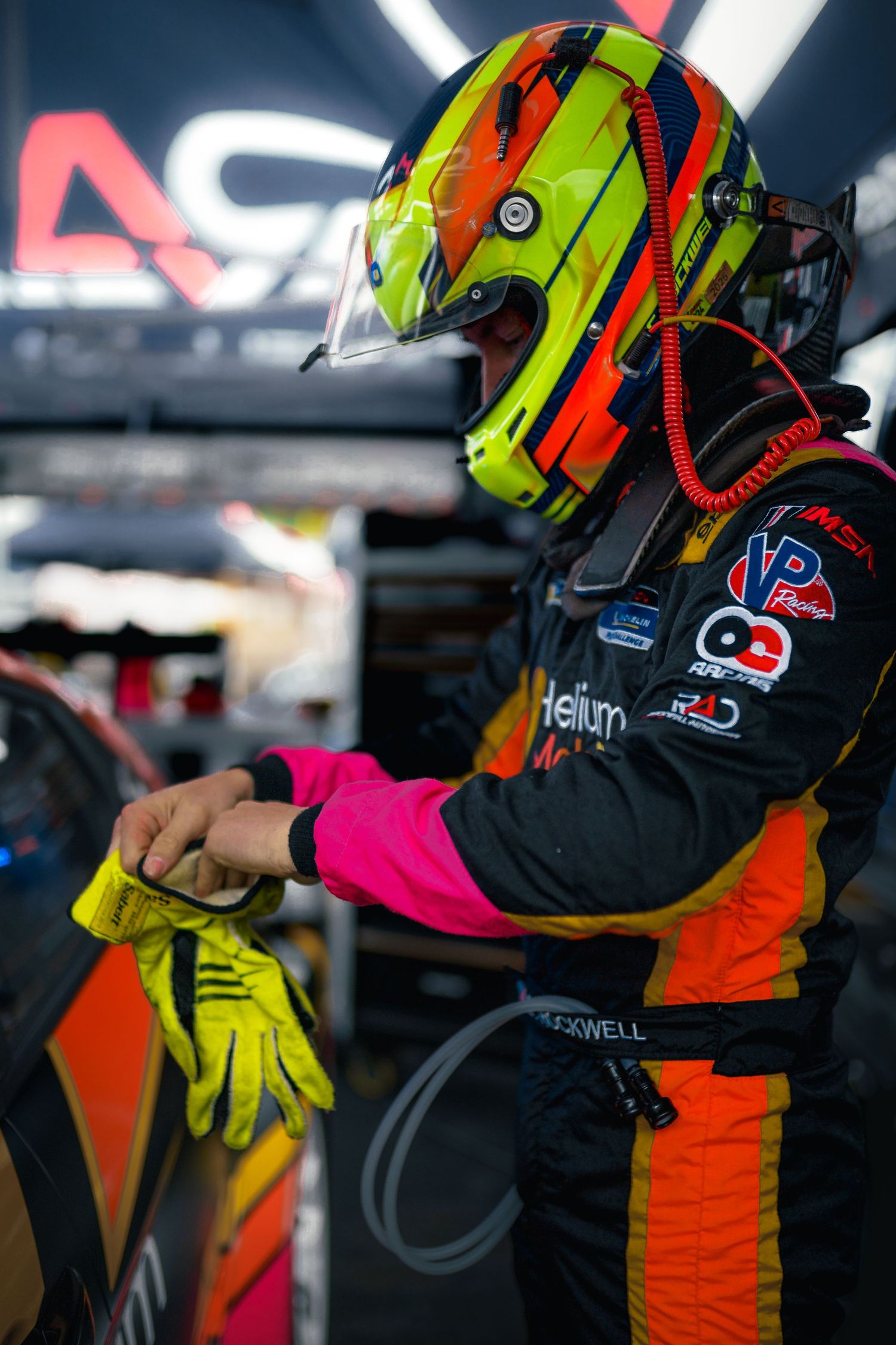 Race car driver adjusts equipment while wearing a neon yellow and orange racing helmet in pit lane.