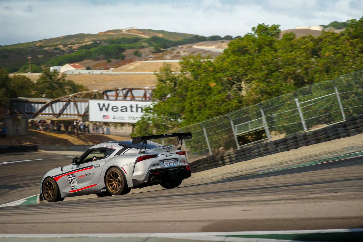 Race cars speed through a winding track at Laguna Seca raceway with rolling hills in the background.