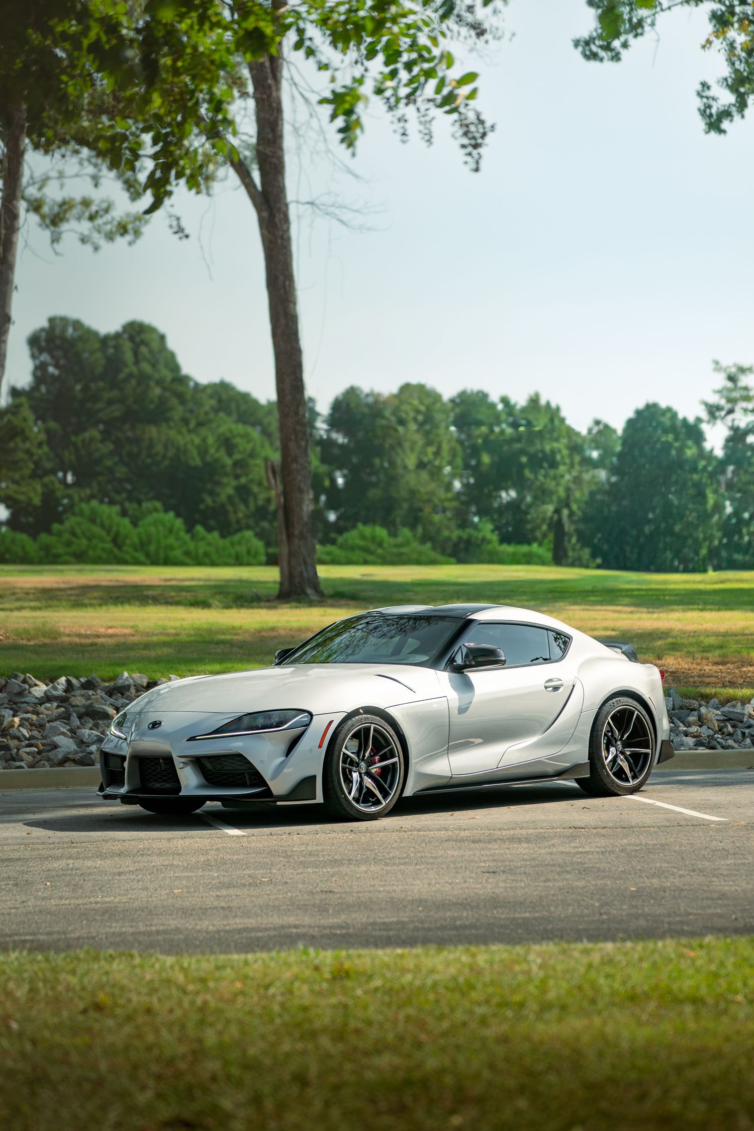 Side profile of a silver Toyota Supra parked on asphalt against a scenic golf course backdrop.