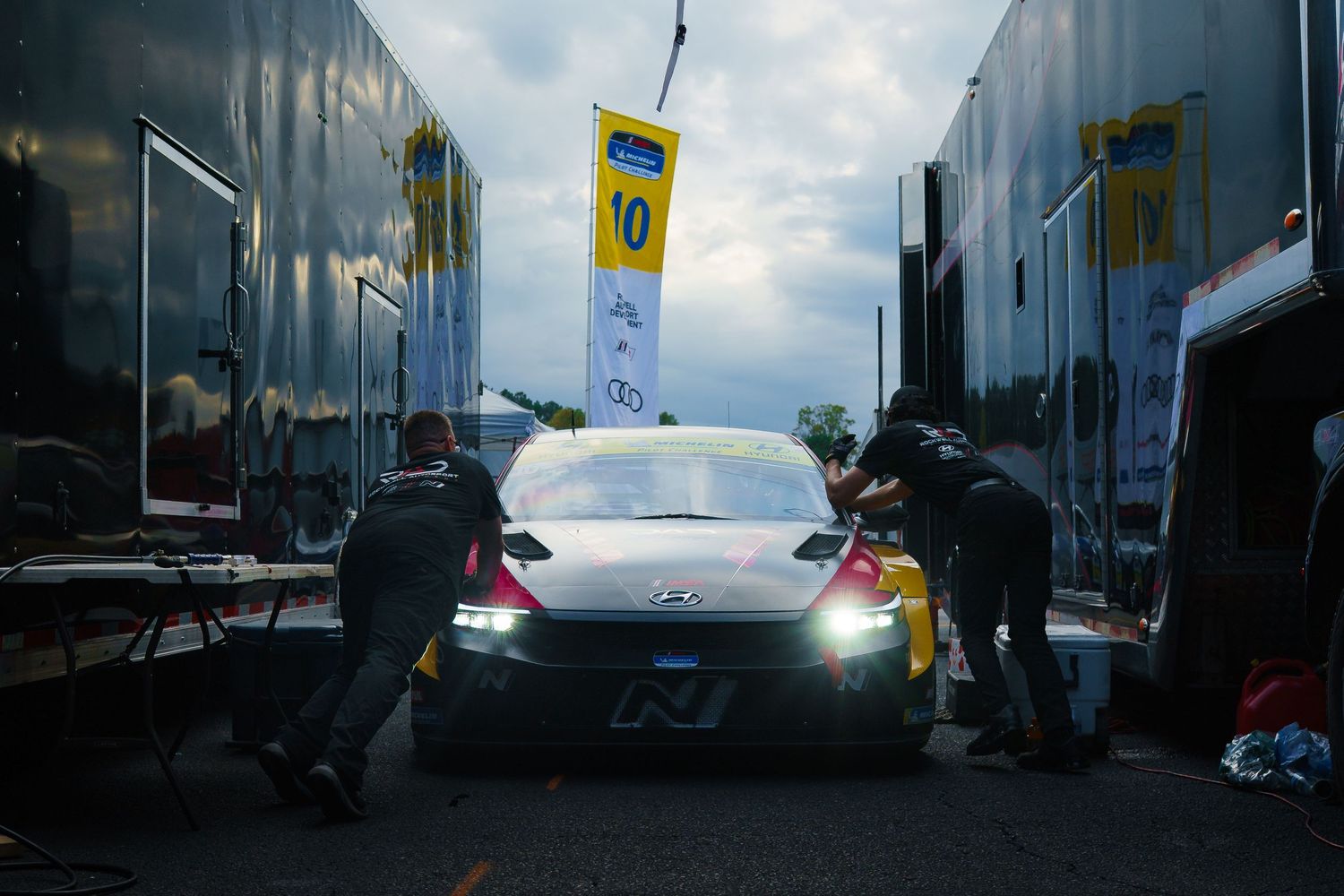 Mechanics work on a race car during pit stop with blue flags and cloudy sky in the background.