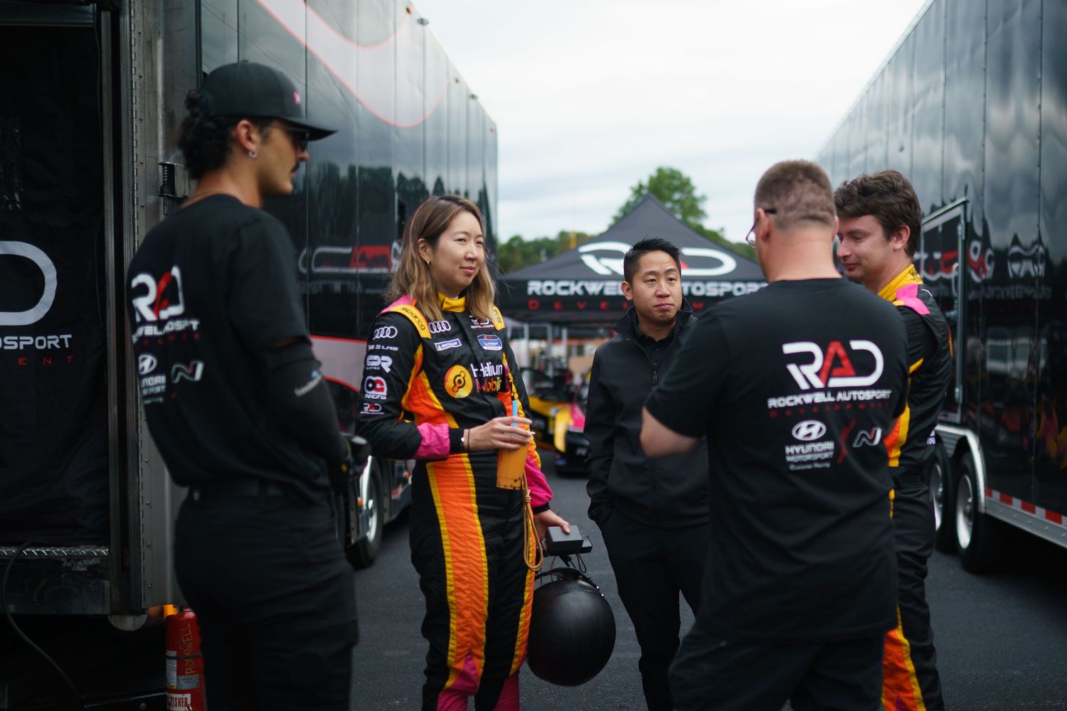 Racing team members gather near transport trucks in the pit area during a motorsport event.