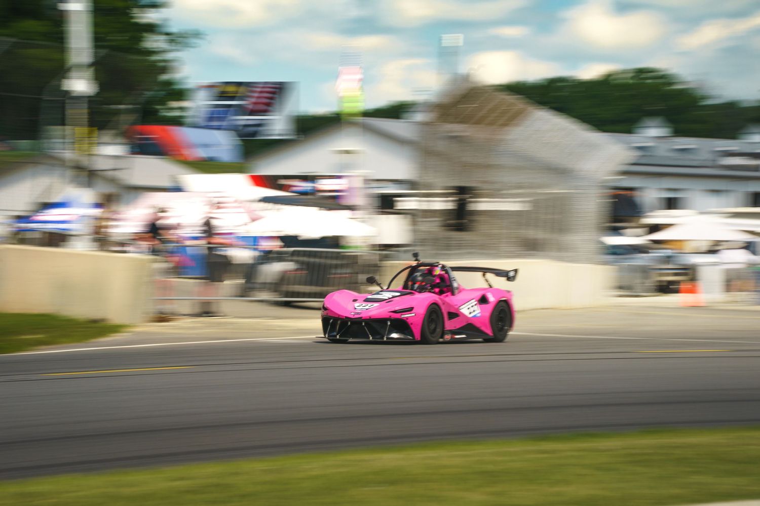 A bright pink race car speeds past on a racetrack with tents and spectators visible in the background.