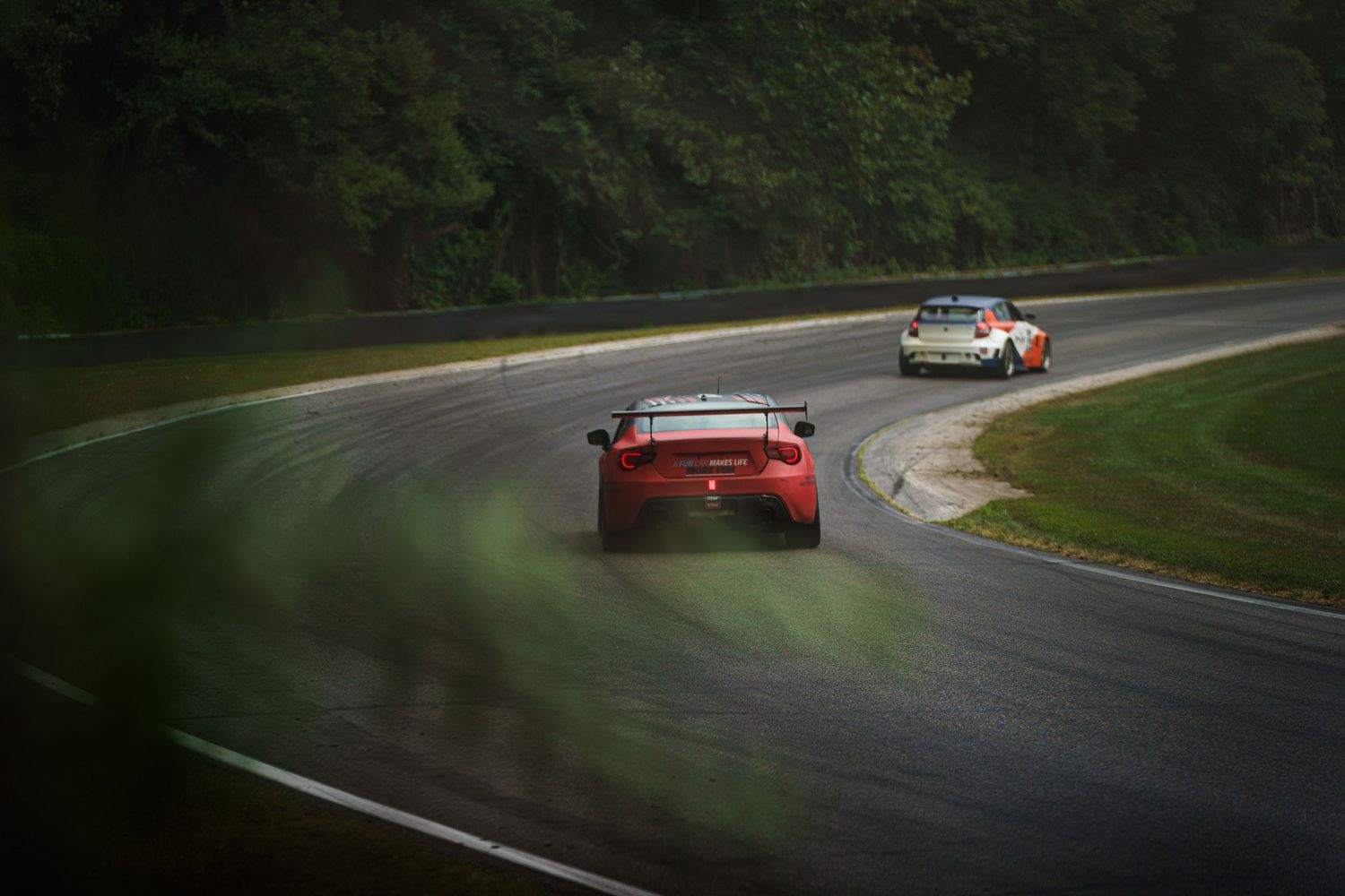 Red racing car follows tight curve on wet racetrack with another vehicle visible ahead during motorsport competition.