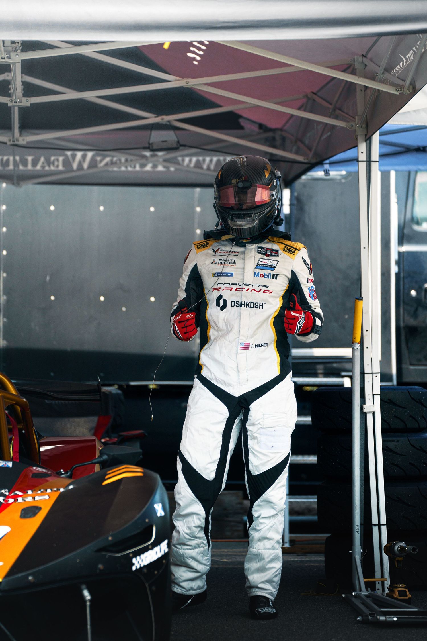 Racing pit crew members in white protective suits work in a garage area during motorsport event preparations.